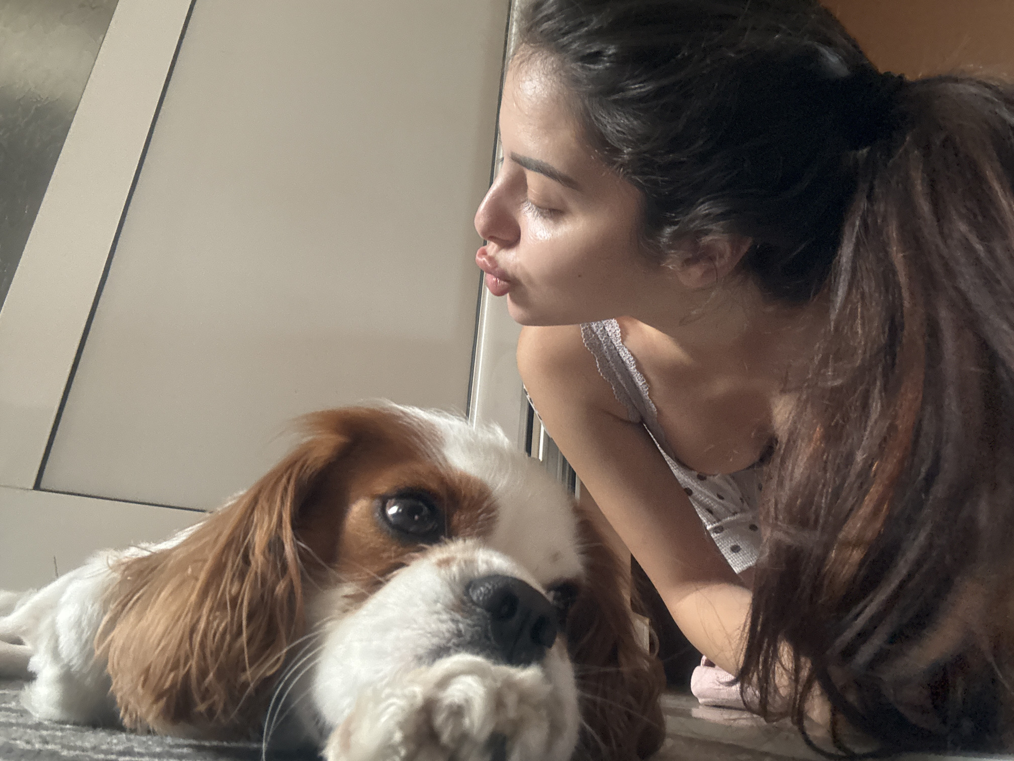 A low-angle indoor photograph captures a tender moment between a young woman and her dog in Plovdiv, Bulgaria. The woman, positioned on the right side of the frame, has dark hair tied back in a ponytail. She is seen in profile, leaning down towards the dog with her eyes closed and lips pursed in a kissing gesture. She wears a light-colored top with small dark polka dots and thin straps, and her skin shows highlights from natural lighting.
In the foreground on the left, a brown and white Cavalier King Charles Spaniel lies calmly on a light grey carpet or rug. Its head rests on its paws, with one dark eye visible, looking towards the viewer. The dog's nose and front paws are slightly out of focus in the immediate foreground. The background consists of light-colored walls or door panels, indicating a domestic setting. The natural, bright light suggests it is daytime. The image conveys a sense of quiet affection and companionship between the woman and her pet. No text is visible.