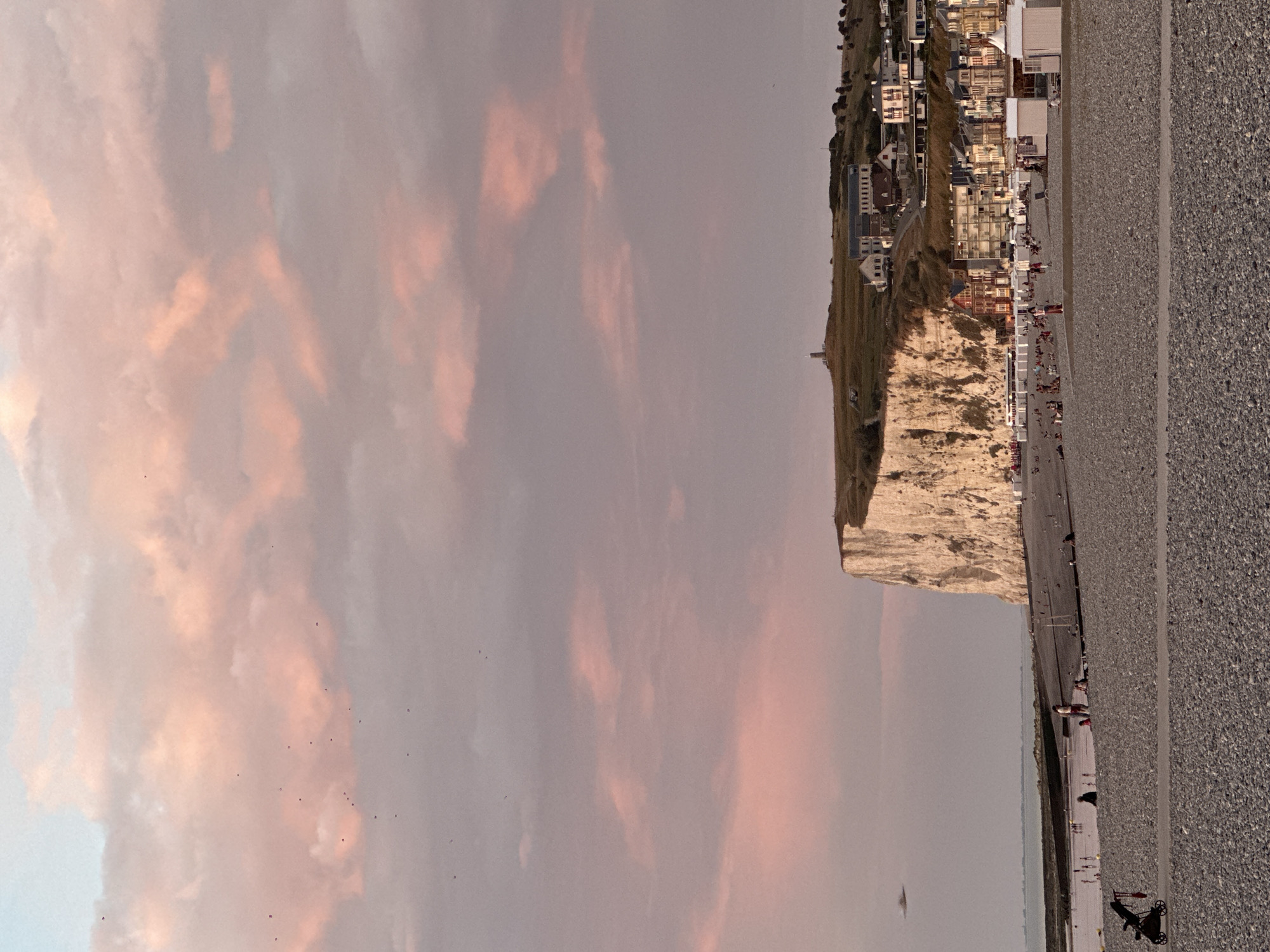 The image depicts a twilight scene at the pebble beach of Le Tréport, France, a seaside resort town in Normandy. In the foreground, the beach is covered in dark grey pebbles. A paved path runs horizontally across the middle ground, with a black baby stroller to the left of the path. Further back, on the beach, there are scattered groups of people, some appearing to be sitting or standing. To the left, a small body of water, likely the sea, is visible.
Dominating the background is a prominent, sheer white cliff face, illuminated by the setting sun, casting a warm glow. Atop the cliff, a cluster of buildings, including houses and a tower, form a small settlement overlooking the sea. Along the base of the cliff, a row of buildings, likely hotels or apartments, faces the beach, with many white beach huts or cabanas lined up in front of them.
The sky is a soft gradient of pale blue and pink, filled with wispy clouds that are tinged with the rosy hues of sunset. Several small, dark specks, likely birds, are visible in the sky. A blurry, unidentified object is present in the sky to the left of the cliff. The overall atmosphere is serene and picturesque, characteristic of a coastal evening.