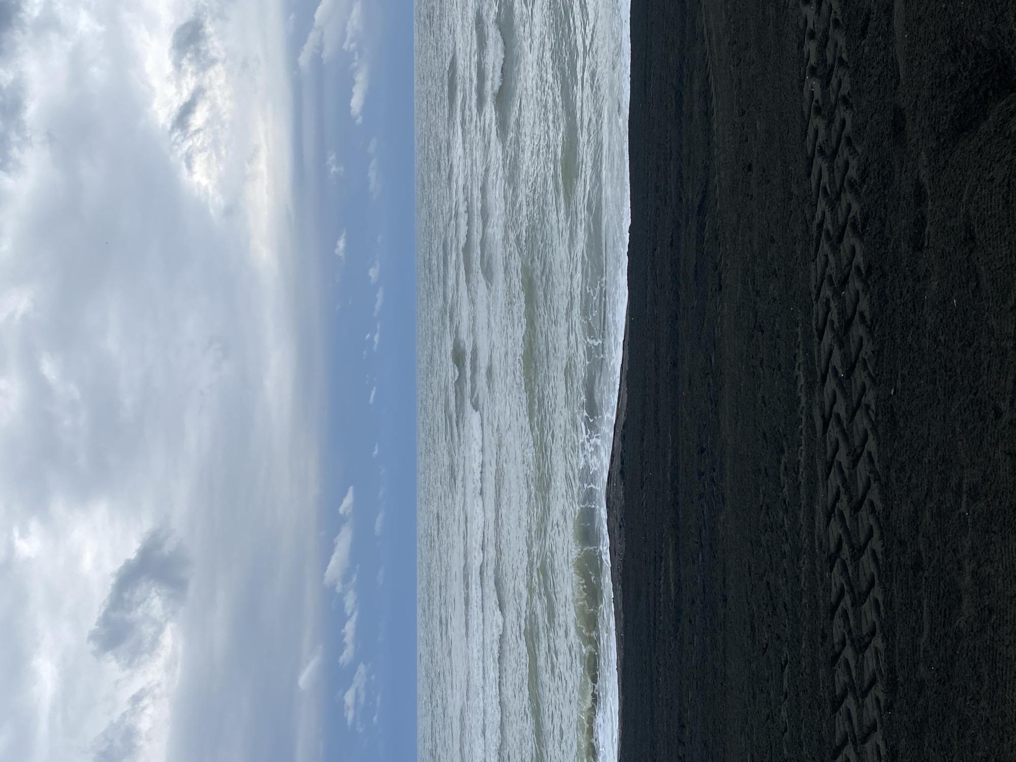 The image depicts a view of the Black Sea from a dark sand beach in Ureki, Georgia. The sky is overcast with grey and white clouds. The sea is a murky grey-green with visible waves and whitecaps near the shore. The beach is composed of fine, black sand, and tire tracks are imprinted on its surface. There are no people or animals visible in the frame. The overall scene conveys a somber and natural coastal environment.