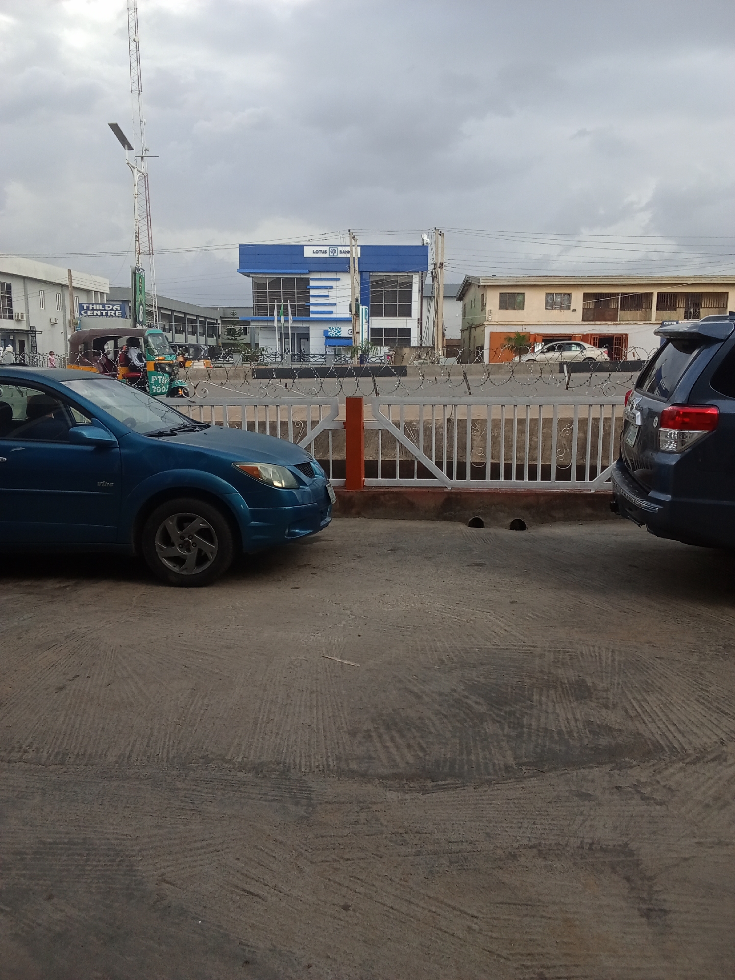 The image depicts a street scene with a blue hatchback car in the foreground, parked on a paved surface. Behind the car, a white metal fence with barbed wire runs horizontally across the scene, separating the road from buildings. Beyond the fence, various structures are visible, including a blue and white building with "LOTUS BANK" signage, and a lighter colored building with multiple windows. Several vehicles, including what appears to be a trishaw, are present in the background. A tall communication mast stands to the left of the frame. The sky is overcast, suggesting cloudy weather. The overall environment appears to be an urban setting, likely within the city of Jos, Nigeria, given the presence of the trishaw and architectural styles. The scene is static, with no prominent actions or interactions observed.