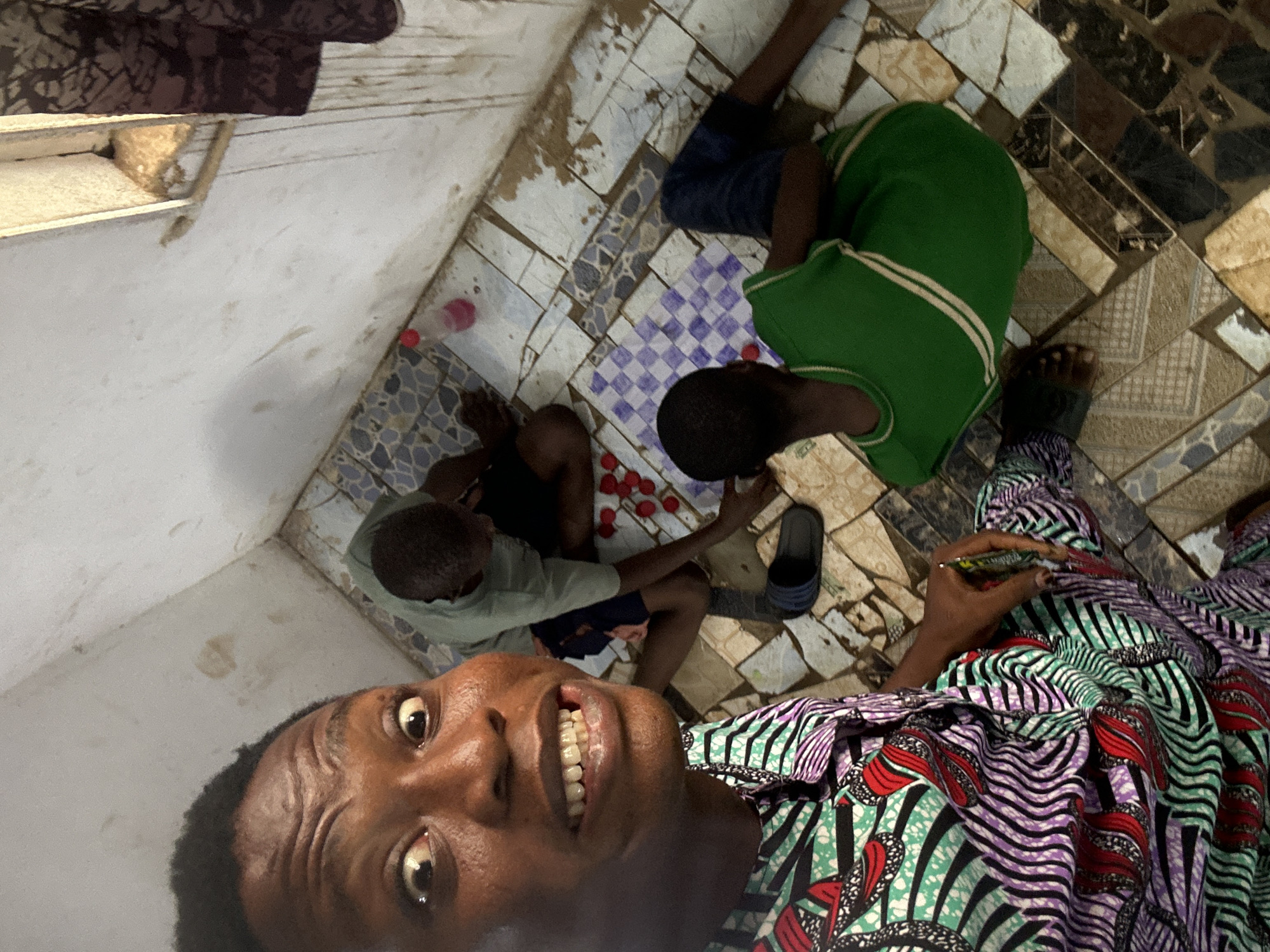 The image depicts three individuals in an indoor setting in the city of Unknown, Unknown. One person is in the foreground taking a selfie, wearing a colorful, patterned top. In the mid-ground, two individuals are kneeling and appear to be engaged in a game or activity involving small, red objects. One person is wearing a green shirt and dark shorts, while the other is wearing a lighter green shirt and patterned shorts. A pair of blue sandals is near the kneeling individuals. The floor is tiled with a patchwork pattern of light and dark tiles, including a section with a blue and white checkerboard pattern. The background shows a white wall and a glimpse of a dark-colored object, possibly furniture, near a window.