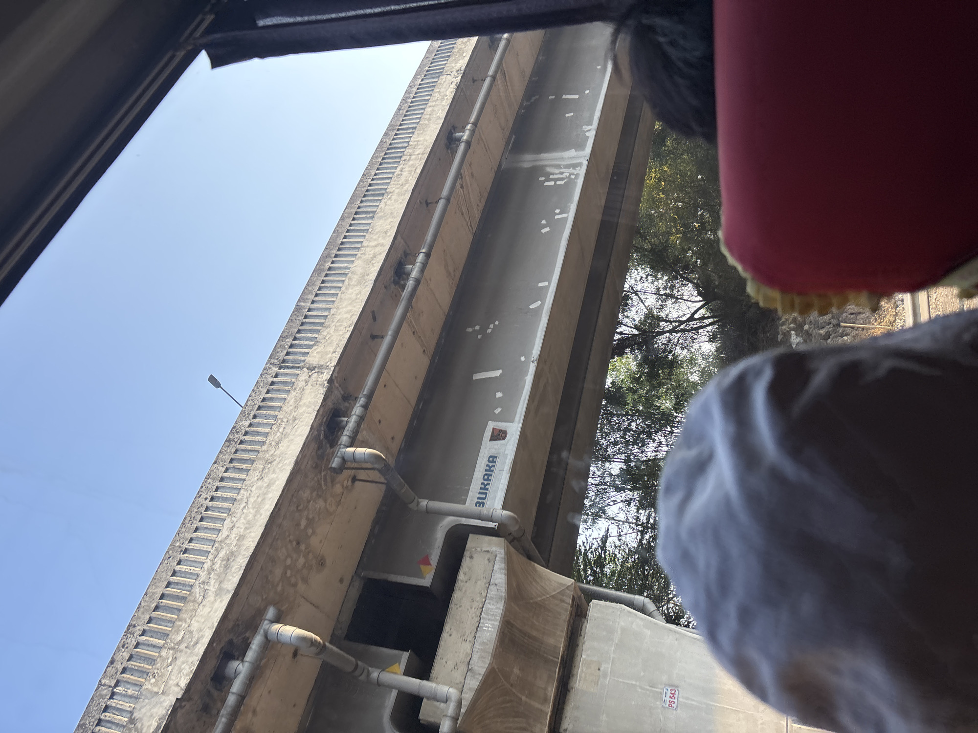 The image, taken from inside a vehicle, shows an elevated concrete structure, likely a bridge or overpass, against a clear blue sky. The structure appears to be part of an infrastructure project in Karawang, Indonesia, as indicated by the "BUKAKA" logo visible on the underside of the bridge. Metal pipes are attached to the underside of the structure. In the foreground, the heads of two passengers are visible, one wearing a white cap and the other a dark, possibly black, hairstyle. Trees and foliage can be seen beneath the elevated structure, suggesting an outdoor, possibly semi-urban or rural, environment. The lighting suggests it is daytime, likely morning or afternoon.