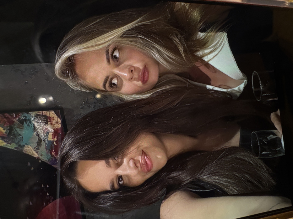 Two young women are reflected in a glossy black surface, possibly a table. The blonde woman at the top has light blonde hair with darker roots and wears a white top. Her eyes are wide, and she appears to be looking at something off-camera. Below her is a woman with long, dark brown hair, smiling slightly with her head tilted. Two empty glasses are visible near the top right corner. A colorful artwork, possibly a painting, can be seen reflected near the bottom left. The overall impression is of a social gathering in a dimly lit place.
