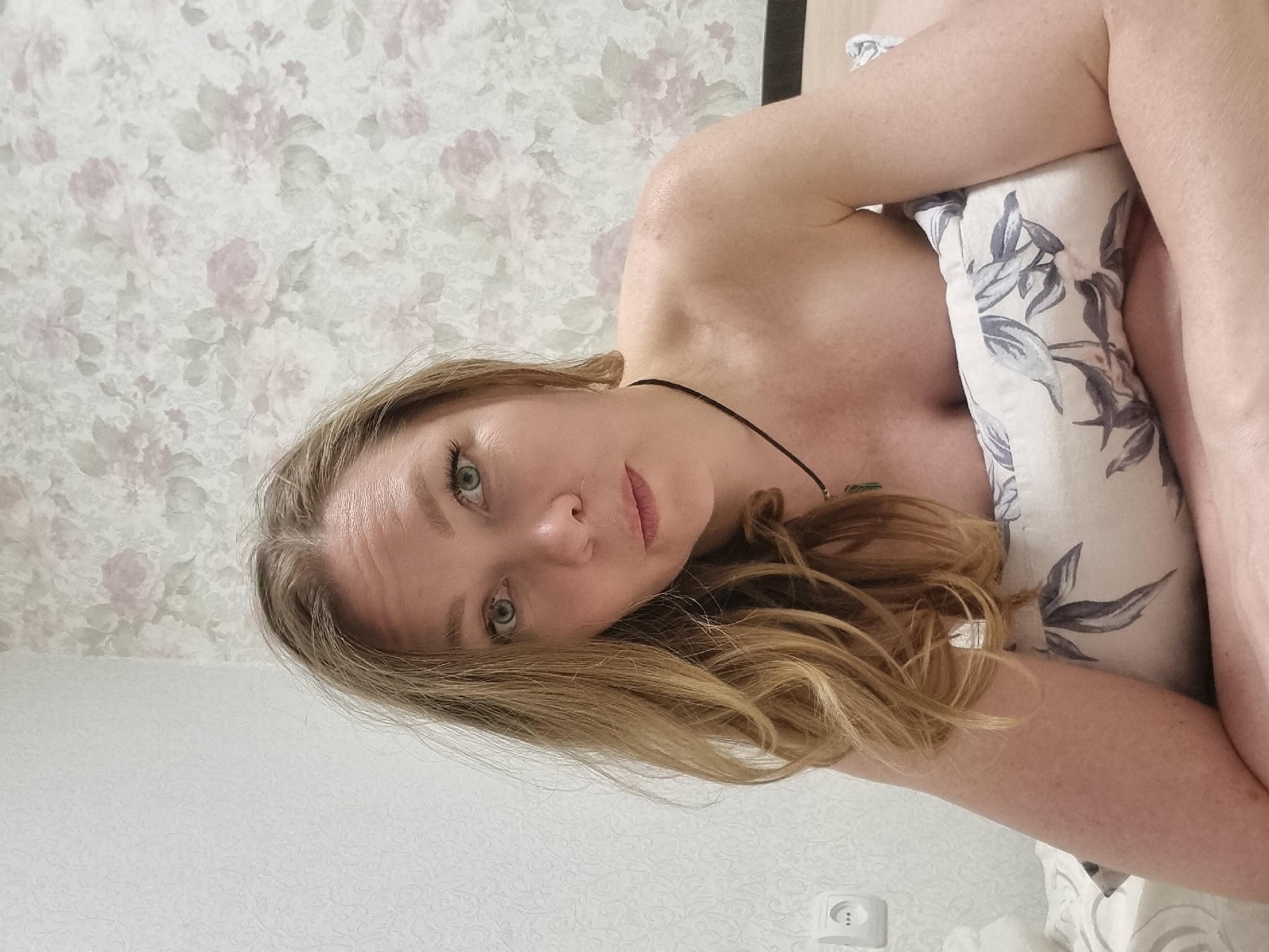 A woman with long, blonde hair and green eyes is lying on a bed, partially covered with a patterned pillow and bedding. She is wearing a thin necklace. The background consists of a floral patterned wallpaper and a plain wall with an electrical outlet. The image appears to be taken in an indoor setting. While the image might be taken in a city like Chaikovsky, Russia, no details in the image confirm this location.