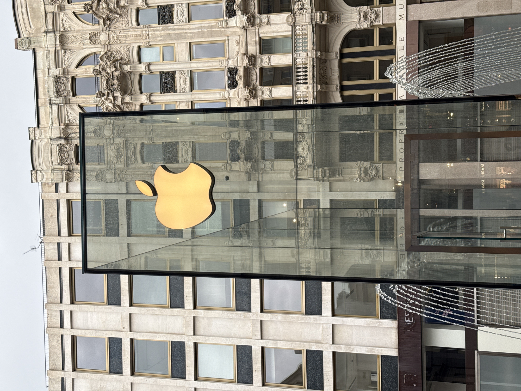 The image displays a prominent, golden Apple logo affixed to a large, clear glass facade, likely part of an Apple Store in an urban setting. The surrounding environment is a striking blend of architectural styles. Reflected and partially visible, an ornate, classical-style building made of light-colored stone features intricate carvings, arched windows, and decorative cornices. Adjacent to this, or reflected on the other side, is a more modern structure characterized by a grid pattern of windows set into darker stone panels.
The scene is static, devoid of human activity, capturing a quiet moment in the city.
The soft, diffused daylight suggests an overcast sky, as seen in the uniform grey background, and there are no harsh shadows. A complex interplay of reflections on the glass facade superimposes and distorts the surrounding architecture, creating a dynamic visual blend of historical grandeur and contemporary design, characteristic of central Milan. A decorative string of white lights, resembling a chandelier or artistic installation, is visible hanging near the right side of the glass facade.
Several pieces of text are discernible in reflection on the glass. These include partial words like "A ALE MU" (possibly part of "MUSEUM") and "PROPRIA" or "PROPR," likely from the ornate building. A reversed reflection also shows "MARI," potentially from an adjacent business name.
