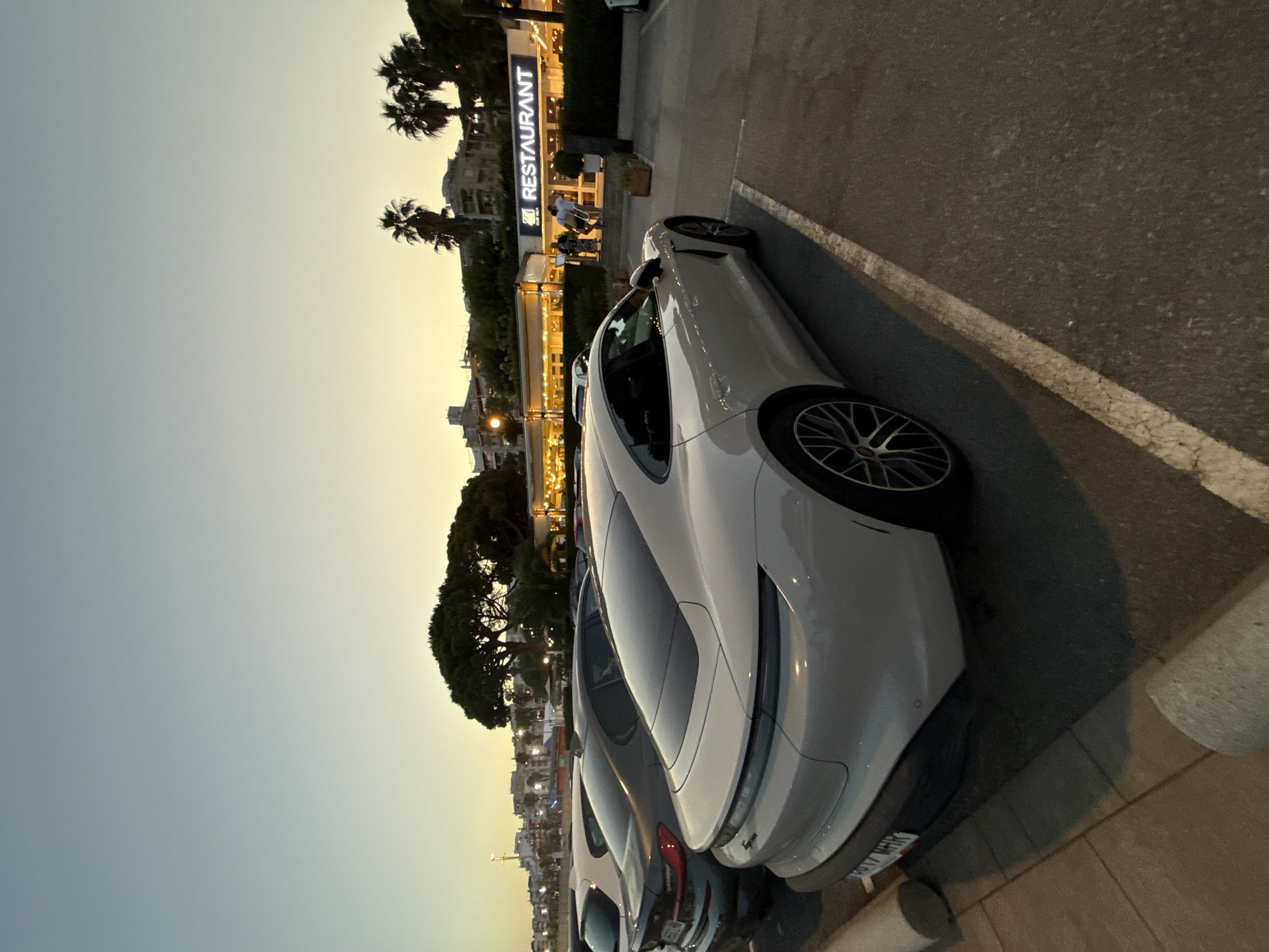 The image depicts a parking lot at dusk in Blanes, Spain, near a restaurant. In the foreground, a white Porsche Taycan is parked, its rear end prominently visible. To its left, a dark-colored BMW sedan is partially visible, parked in an adjacent spot. The background features a building with a large sign that reads "RESTAURANT," illuminated by warm interior lights. Several palm trees and other foliage are scattered throughout the scene, and a few people are seen standing near the entrance of the restaurant. The sky displays a gradient of soft blues and yellows, characteristic of twilight.