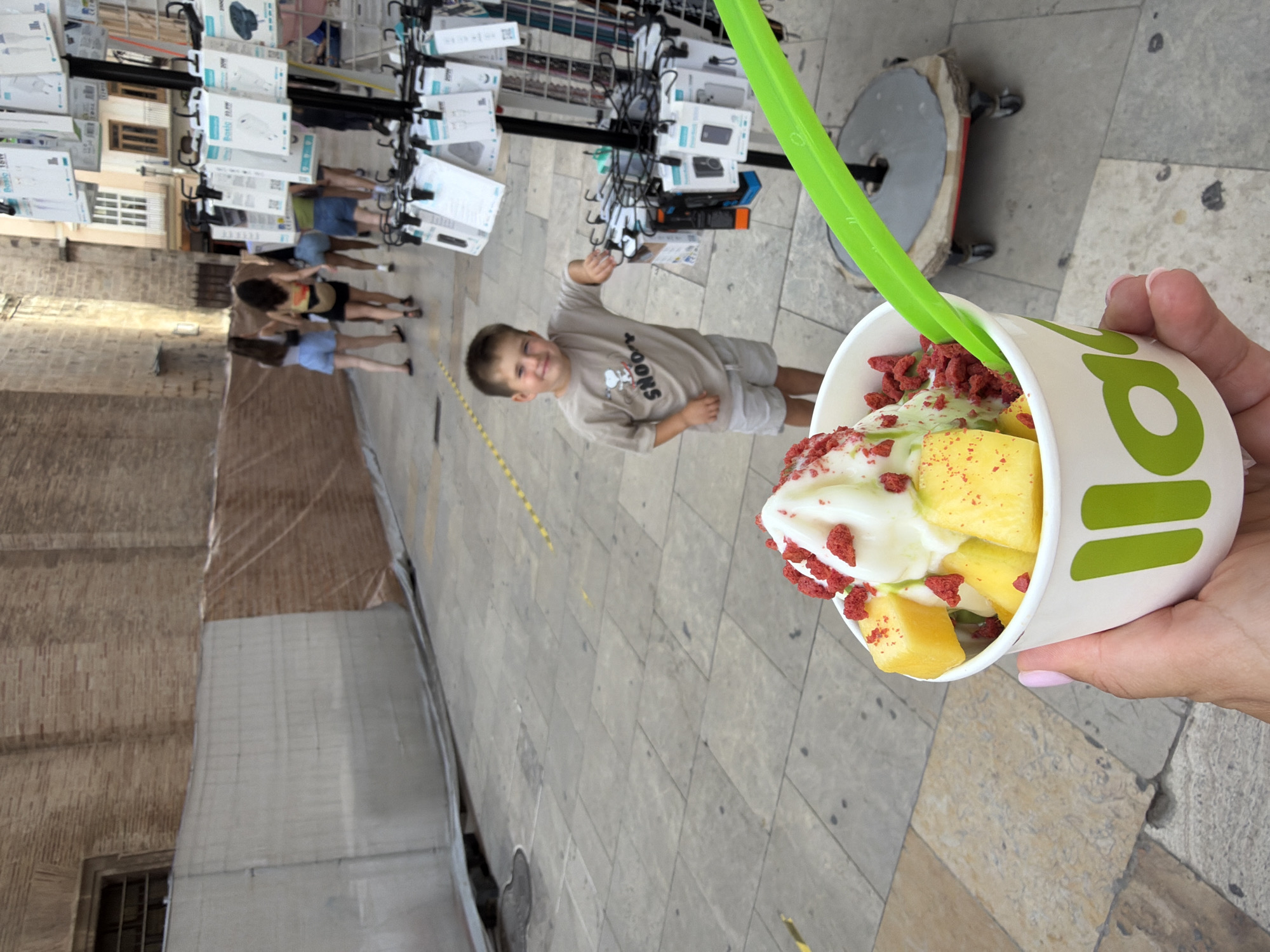 In an outdoor urban setting with stone-paved ground, a person's hand holds a white cup of ice cream topped with cubed yellow fruit and red sprinkles. A green spoon is placed in the ice cream. A young boy in a tan t-shirt and grey shorts stands in the middle ground, smiling and leaning on a display rack filled with packaged phone accessories. Several young women are visible walking in the background. A brick building is on the left side of the image, and a temporary barrier covered with netting and tarpaulin is present.
