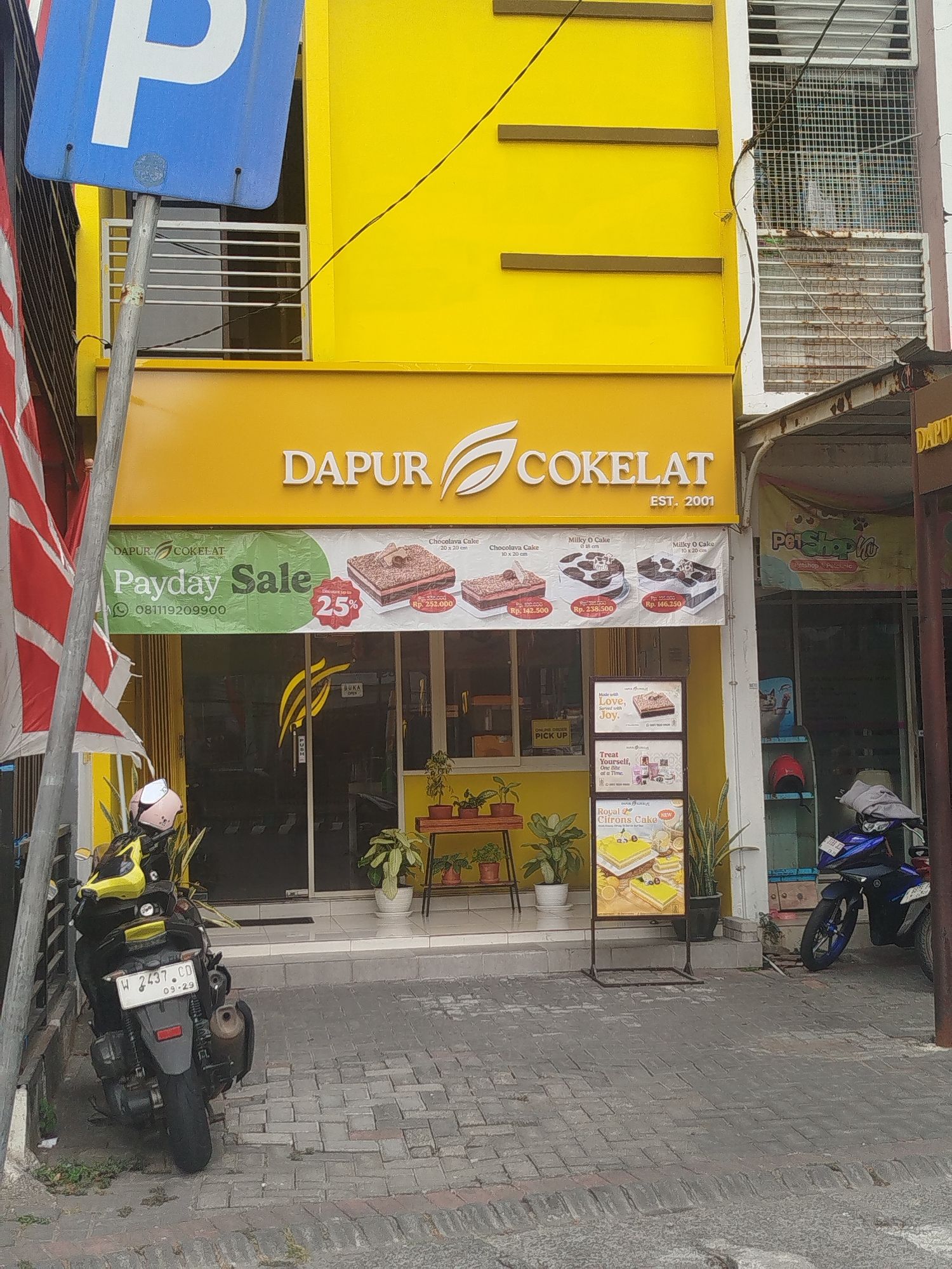 The image shows the exterior of a "Dapur Cokelat" (Chocolate Kitchen) store located in Yosowilangun, Indonesia. The building is painted bright yellow and features a prominent "DAPUR COKELAT" logo with a stylized chocolate swirl. A banner for a "Payday Sale" is displayed, advertising up to a 25% discount on cakes. Several cake images, including a "Chocolava Cake" and "Milky O Cake," are visible on the banner with their prices.
In front of the store, a dark-colored motorcycle with a white license plate (W 2437 CD) is parked on a patterned brick sidewalk. A person wearing a light pink helmet is seated on the motorcycle. To the left of the store, a blue parking sign with a white "P" is mounted on a metal pole, partially obscuring a striped flag. Various potted plants are placed near the entrance. A display stand showcases three framed advertisements for different cakes. Further to the right, a "Pet Shop" sign is visible, suggesting a neighboring business. The overall environment appears to be an urban street scene.