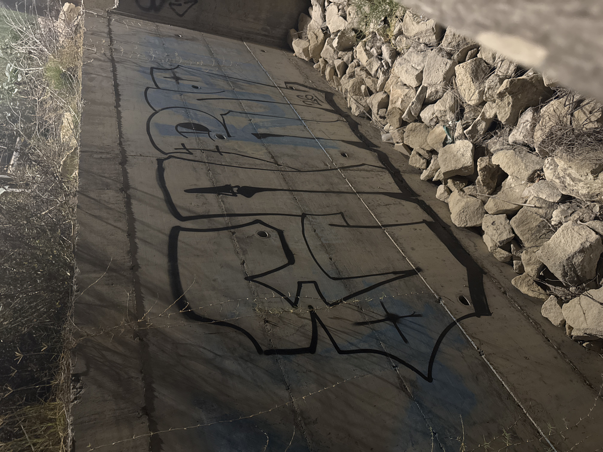 The image shows a graffiti-covered concrete wall, likely part of an urban drainage channel or a similar infrastructure, located in Agios Tychona Community, Cyprus. The graffiti is in black with some blue spray paint visible underneath. The wall is weathered and has a series of horizontal lines or joints. There are several small holes in the concrete. At the base of the wall, a pile of rocks and dry vegetation is visible, indicating an outdoor, possibly neglected, area. The lighting suggests it is nighttime or a dimly lit environment. The graffiti appears to be stylized lettering, and a small tag with the number "26" is visible on the wall.