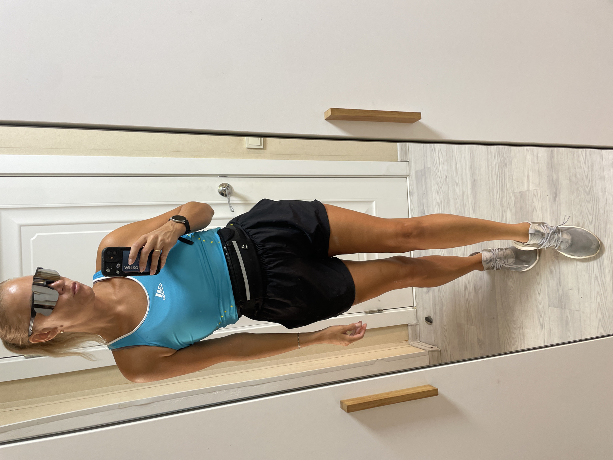A blonde woman stands in front of a mirror, taking a selfie. She is wearing a blue tank top with an Adidas logo, black athletic shorts, a black fanny pack, and grey sneakers. She has on large, reflective sunglasses and her hair is pulled back in a ponytail. The background appears to be an indoor setting, possibly a home or a gym, with a white door and a wooden closet handle visible. There is no indication that the image was taken in Baranovka, Russia.