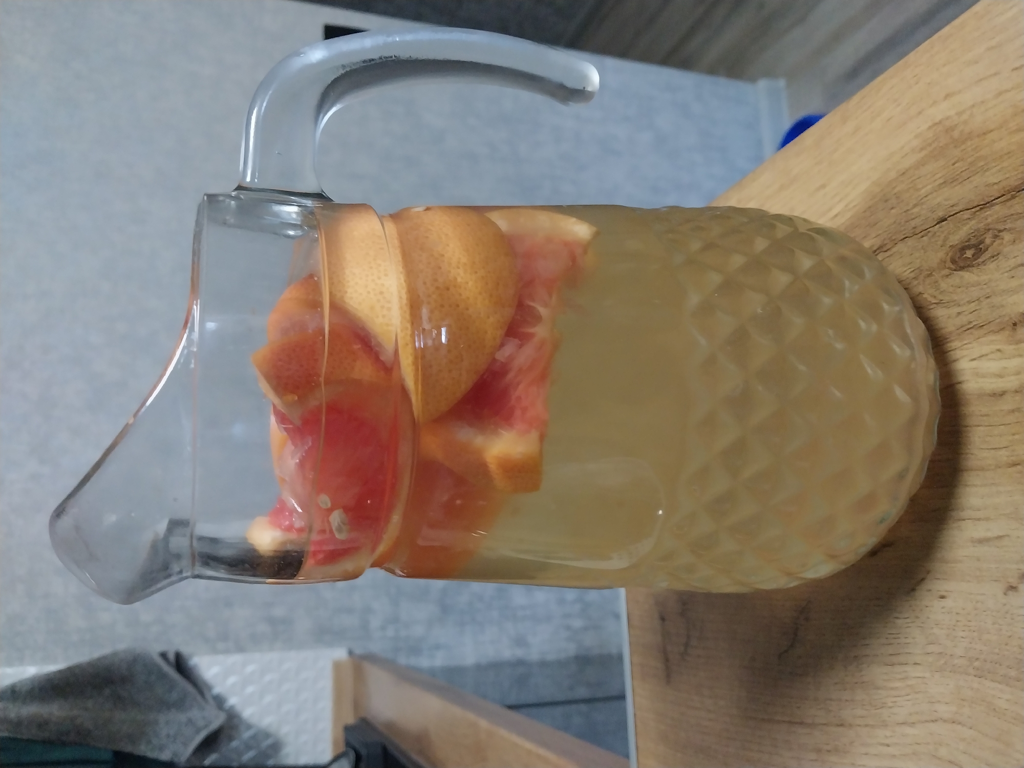 A glass pitcher filled with a pale yellow liquid and slices of grapefruit is positioned on a wooden surface. The pitcher has a diamond-shaped pattern on its lower half and a large, curved handle. In the background, a gray towel hangs on the left, and a wall with light gray textured wallpaper and a darker gray floor is visible. The scene appears to be in an indoor setting, likely a kitchen or dining area. There are no people or animals present. The lighting is soft and diffused, suggesting it may be daylight or indoor lighting. The image is taken from a slightly low angle, looking up at the pitcher.