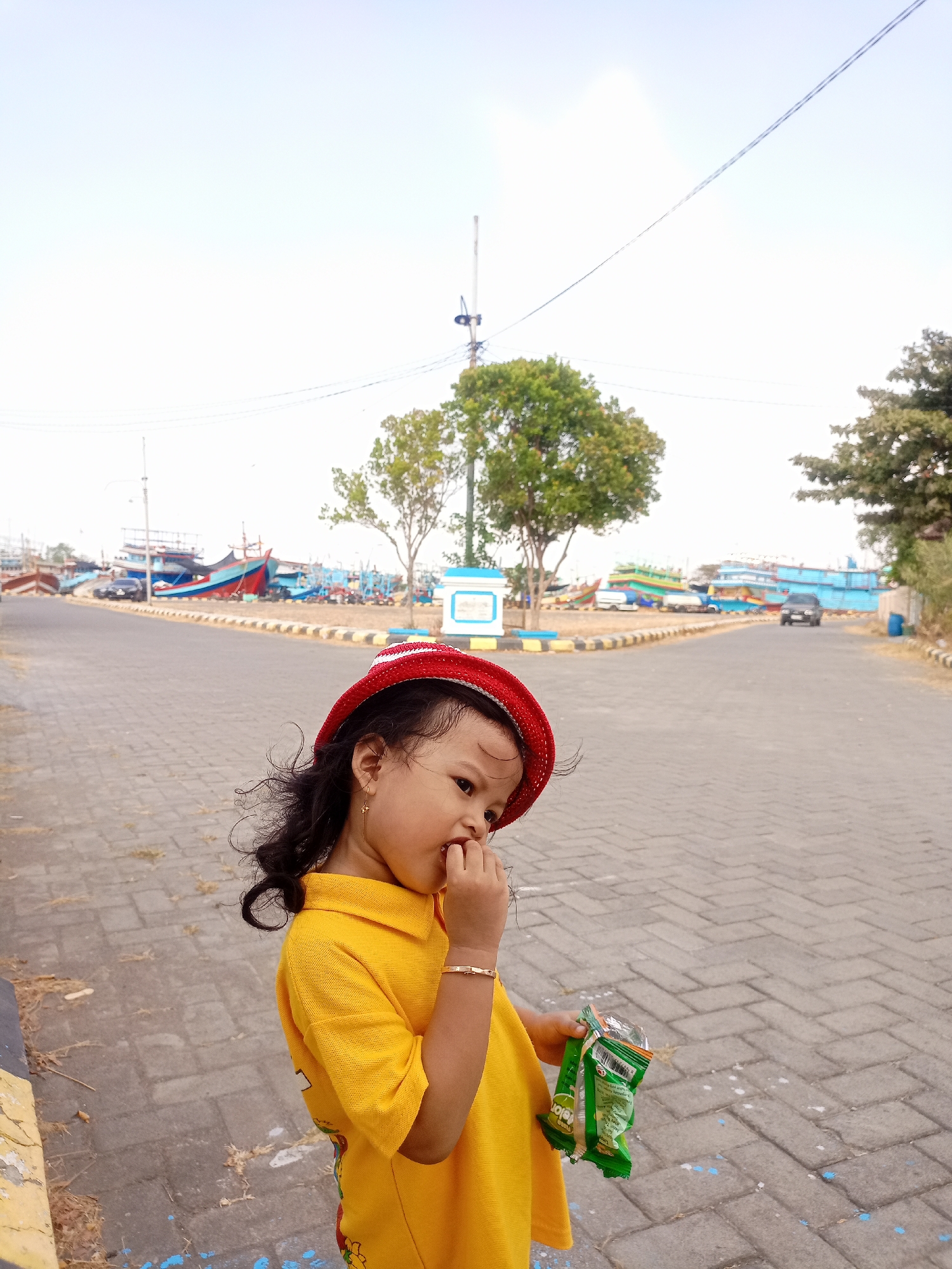 A young girl wearing a red hat and a yellow shirt is standing on a paved walkway. She has dark curly hair and is holding a green bag of snacks in her right hand while bringing her fingers to her mouth. In the background, there is a road with parked cars and boats docked at a port. The scene appears to be in the city of Juwana, Indonesia, given the presence of the harbor and fishing boats, which are characteristic of Juwana's maritime activities. The sky is bright and overcast.