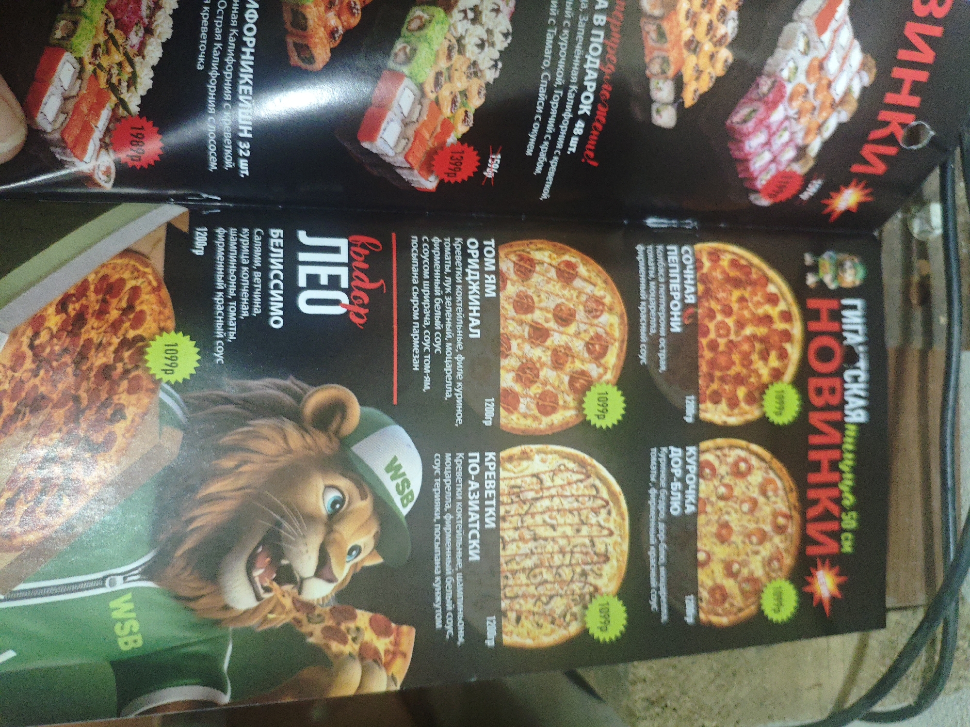 This image appears to be a close-up, slightly angled shot of a menu for a food establishment, likely a restaurant or cafe. The menu features vibrant images of various food items, primarily pizzas and sushi rolls. The text is in Russian.
On the left side of the image, a large pepperoni pizza is prominently displayed, with a cartoon lion character wearing a green and white baseball cap that says "WSB" holding a slice of pizza. Below the lion, another pizza is partially visible.
In the upper portion of the menu, there are images of sushi rolls, with prices indicated such as "1989p" and "1399p". Text in Russian describes the sushi options.
Further to the right, several more pizzas are showcased. One is labeled "ТОМ ЯМ ОРИДЖИНАЛ", another "КРЕВЕТКИ ПО-АЗИАТСКИ", and a third "КУРОЧКА ДОР-БЛЮ". These also have accompanying Russian descriptions and prices like "1099p" and "1200p".
The overall aesthetic is colorful and appetizing, designed to attract customers. The quality of the image suggests it was taken with a smartphone or similar device, possibly in low lighting conditions given the shadows and some blur. There is no visible indication of the specific location within Armavir, Russia, beyond the fact that it is a menu from an establishment there.