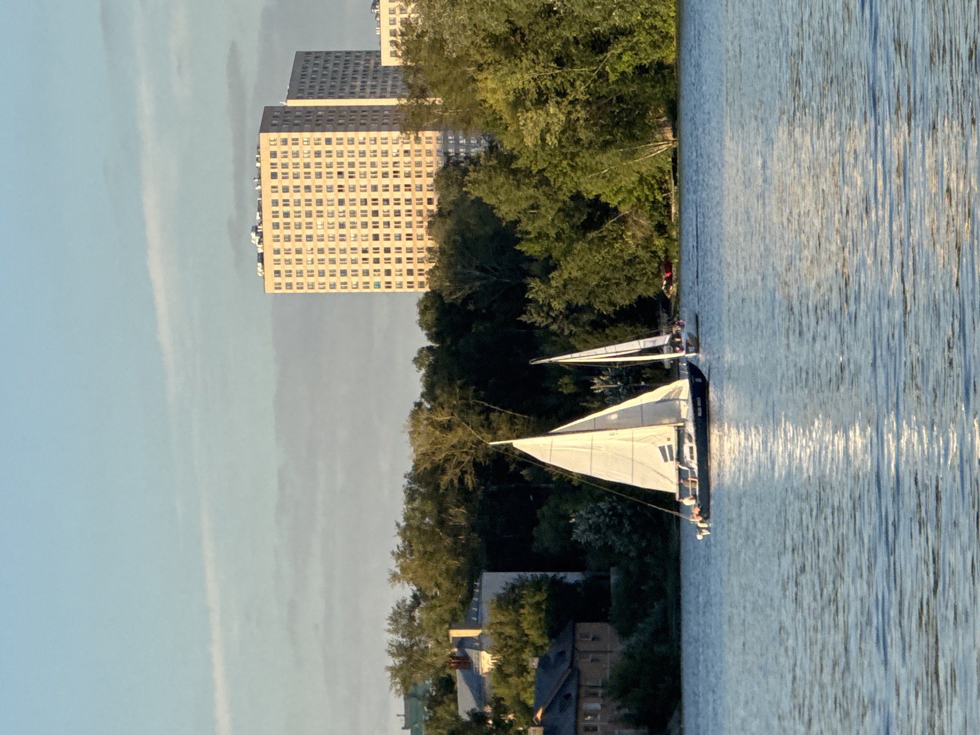 The image depicts a scenic view of the Khimki Reservoir in Khimki, Russia, bathed in the soft, warm light of a setting sun. In the foreground, the calm, blue water of the reservoir reflects the sky and the surrounding landscape. Two sailboats are prominently featured on the water. The larger sailboat, on the left, has its white sail fully deployed, displaying a dark hull and what appears to be a few people on board. A second, smaller sailboat is visible further to the right, also with its sail up.
The background reveals a lush green treeline that separates the water from the urban environment. Beyond the trees, a row of apartment buildings rises into the sky. Two tall, light-colored residential buildings are the most striking structures, with numerous windows clearly visible. To the left of these modern buildings, older, lower-rise houses with red roofs are nestled amongst the trees. The sky above is a pale blue with wispy clouds, indicating a pleasant evening. The overall impression is one of peaceful coexistence between nature and the urban setting.