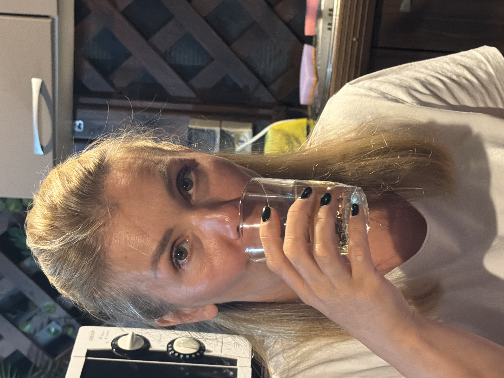 A close-up, medium shot shows a fair-skinned Caucasian woman with blonde hair pulled back, wearing a white t-shirt, as she drinks from a clear glass. She is holding the glass with her right hand, her index finger extended and her thumb bent. Her fingernails are painted black. The woman's face is turned slightly to her left, and her eyes are open and looking forward. The background appears to be an outdoor kitchen or patio area with a dark lattice-patterned screen, a microwave oven, and cabinets. The overall lighting suggests it is daytime. The location of Sochi, Russia, is not discernible from the visual information in the image.