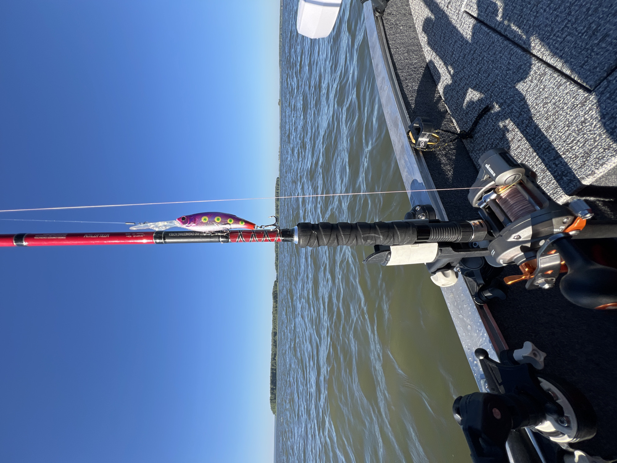 An eye-level, medium shot captures fishing gear on a boat on a body of water, with a distant treeline on a calm, sunny day. The foreground is dominated by a fishing rod with a purple and white lure attached, held in a black rod holder on the boat's gray, textured deck. To the left of the rod holder are two other fishing reels, one black with silver accents and the other silver with orange and black accents. The water, which is a murky green, has small ripples and reflects the blue sky. In the distance, a long, low treeline with a few smaller islands is visible on the horizon. The sky is a clear, cloudless blue. The scene is set in Novopichugovo, Russia.