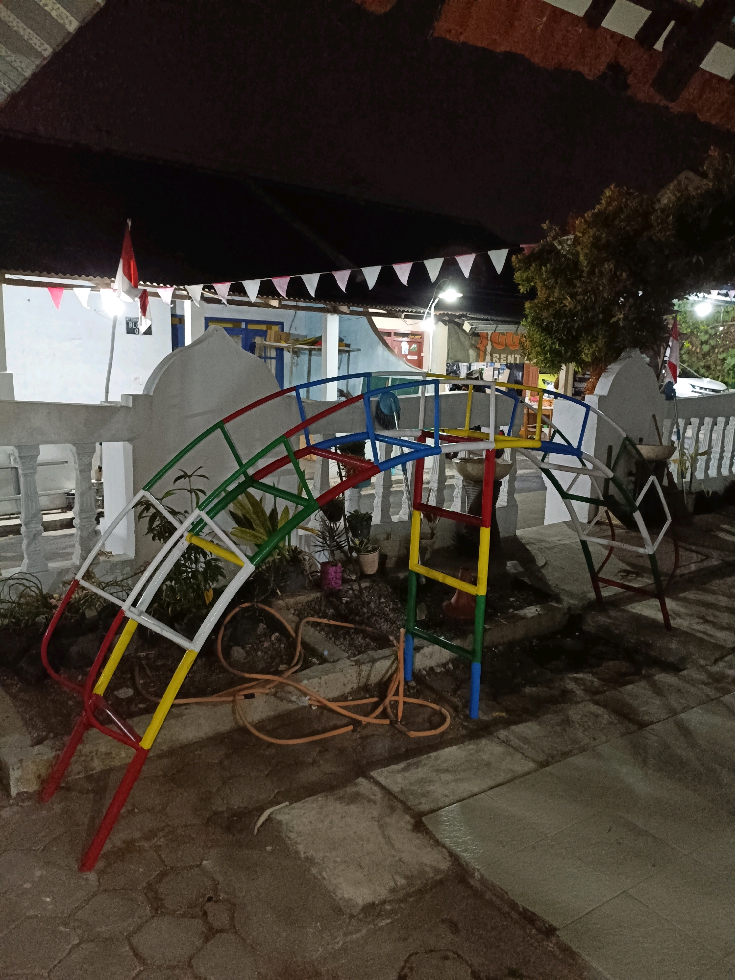 The image depicts an outdoor scene in Maguwoharjo, Indonesia, at dusk or night, with artificial lighting illuminating the area. In the foreground, a colorful metal playground structure, painted red, yellow, green, and blue, arches across the frame. The structure appears to be a climbing frame or monkey bars. The ground beneath it is a mixture of soil, gravel, and paving stones. A coiled orange hose is visible on the ground near the structure.
Behind the playground, a white railing with decorative balusters runs along a raised platform. A white, domed structure is visible behind the railing, and further back, a building with a dark, textured roof is apparent. Decorative triangular flags, white and pink, are strung along the edge of the roof. An Indonesian flag, red and white, is also present, mounted on a pole near the building.
Several potted plants are scattered around the area, some appearing to be small shrubs or ornamental plants. In the background, a tree with dense foliage is visible. The overall environment suggests a public space, possibly a park, schoolyard, or community area, in a residential or commercial part of Maguwoharjo. The lighting conditions indicate it is after sunset. There are no people visible in the image.