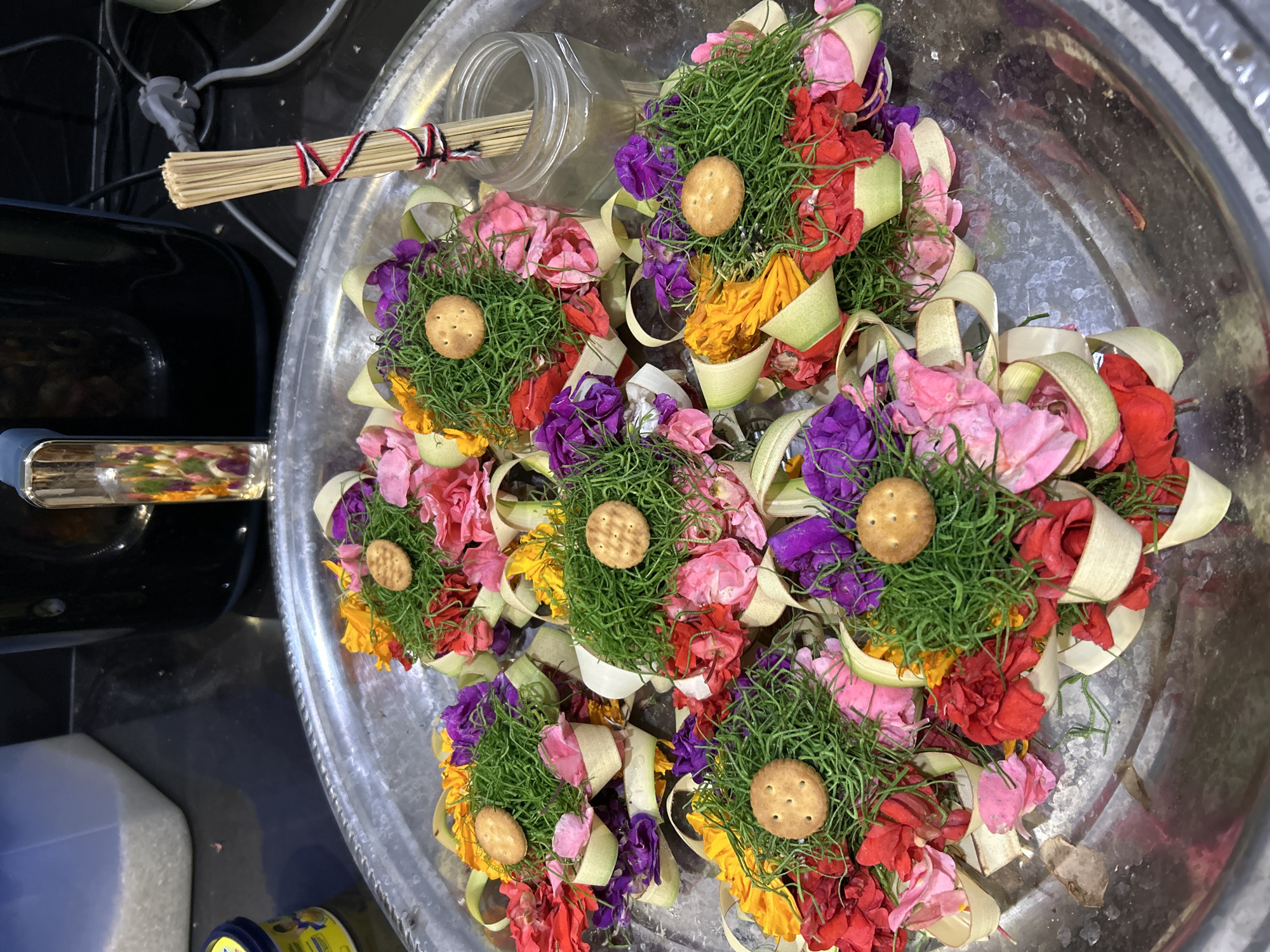 The image shows a close-up, overhead view of several offerings arranged on a large, circular, metal tray. The offerings are small, cone-shaped bundles of vibrant flowers in shades of pink, red, purple, and yellow, interspersed with green grass and delicate, light-colored leaves. Each offering is topped with a single, round, light-brown cracker, creating a contrast in texture and color. The offerings are artfully arranged on the tray, some overlapping each other.
To the left of the tray, a bundle of thin, straw-like sticks is tied with red, white, and black string. Behind the offerings, a clear plastic jar with a wide opening is visible, partially filled with what appears to be similar straw-like sticks and possibly some dried flowers. The tray itself is positioned on what looks like a dark, possibly black, surface, with a metallic handle partially visible on the far left. The overall setting appears to be indoors, possibly in a market or a place of worship, given the nature of the offerings. The lighting is bright, illuminating the colors of the flowers and the reflective surface of the tray. The image captures a moment of preparation or presentation of these ceremonial items. There is no visible text in the image.
