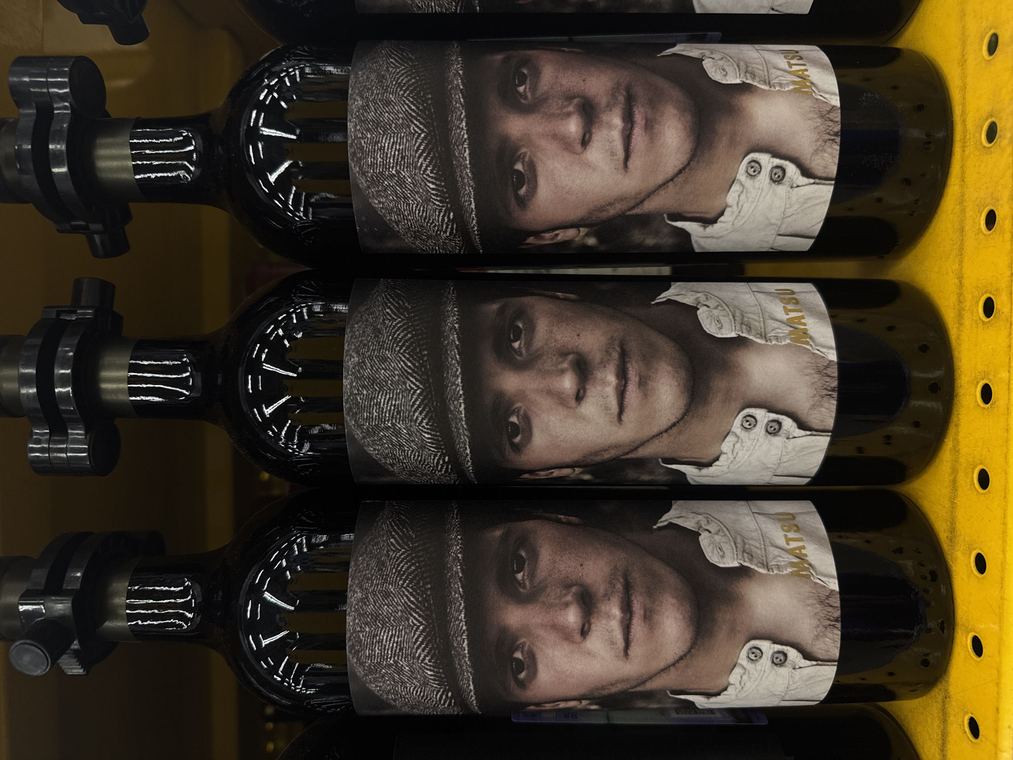 Three bottles of Matsu red wine are stacked in a yellow shelving unit. The labels on the bottles depict a sepia-toned portrait of a man wearing a tweed cap and a partially unbuttoned shirt. The word "MATSU" is printed on the shirt collar of the label. The bottles are in a dark glass, with metal mechanisms attached to the bottle necks.
The location in Saint Petersburg, Russia is not evident in the image.