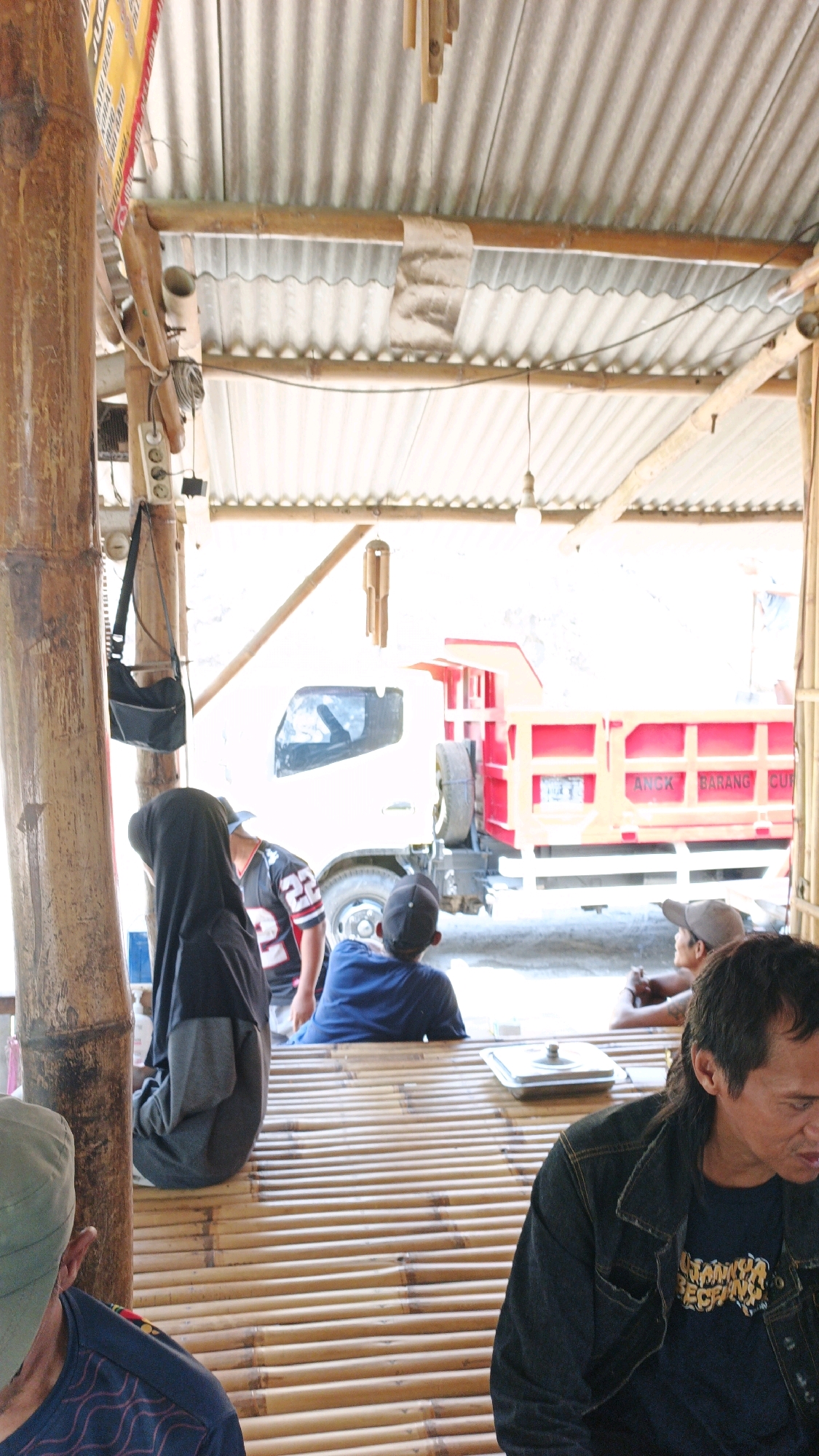 The image depicts an outdoor scene under a corrugated metal roof supported by bamboo poles, likely in Sanggrahan, Indonesia. Several people are present. In the foreground, to the left, a person wearing a green cap is partially visible from the back, and next to them, a person in a black hijab and grey garment is seated. In the middle ground, a person wearing a black jersey with the number "22" is standing, facing away from the camera. To their right, a person in a blue shirt and black cap is seated. Further to the right, a person in a grey cap is seated, facing left. A man in a dark denim jacket and a dark t-shirt with yellow lettering is seated in the foreground on the right, looking down and to the left. A white dump truck with a red bed is visible in the background. The ground appears to be paved or dirt, and a bamboo table is positioned in the foreground. There are electrical outlets and hanging bags on the bamboo pole on the left. Bamboo chimes are hanging from the roof structure.