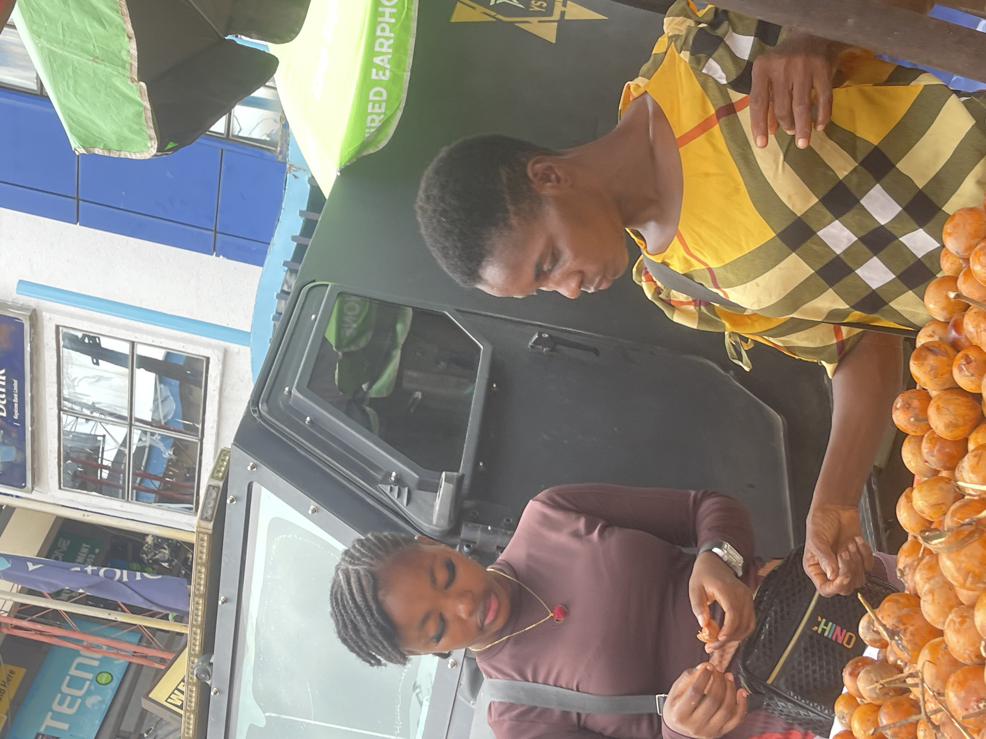 This image, rotated 90 degrees clockwise, captures a vibrant street scene in what appears to be a market or commercial area in Onitsha, Nigeria, during daylight hours.
The primary subjects are two women. The woman on the right, likely a vendor, wears a yellow, black, and white plaid top and has short, dark hair. She stands behind a large display of numerous round, orange-brown fruits, possibly African star apples (agbalumo), which are skewered on sticks. Her hands are visible near the fruits, suggesting she is attending to her stall.
The second woman, a customer, is positioned slightly below and to the left of the vendor. She wears a maroon or brown long-sleeved top and has braided hair. Her attention is focused on a fruit she holds in her hands, appearing to be peeling or preparing to eat it. She wears a gold chain with a red pendant and a watch on her left wrist. A black handbag with "CHINO" in colorful letters is slung over her shoulder.
In the background, a large, dark-colored vehicle, possibly an armored truck or robust SUV, dominates the space between the women. Above and around the scene, two green canopies or umbrellas are visible, one partially covering text that reads "WIRED EARPHO" (likely "WIRED EARPHONE"). Further in the background, various urban elements are visible, including parts of buildings, windows reflecting the surroundings, and signs with partial text like "TECNO" and "Bank." The overall impression is a bustling outdoor commerce setting.