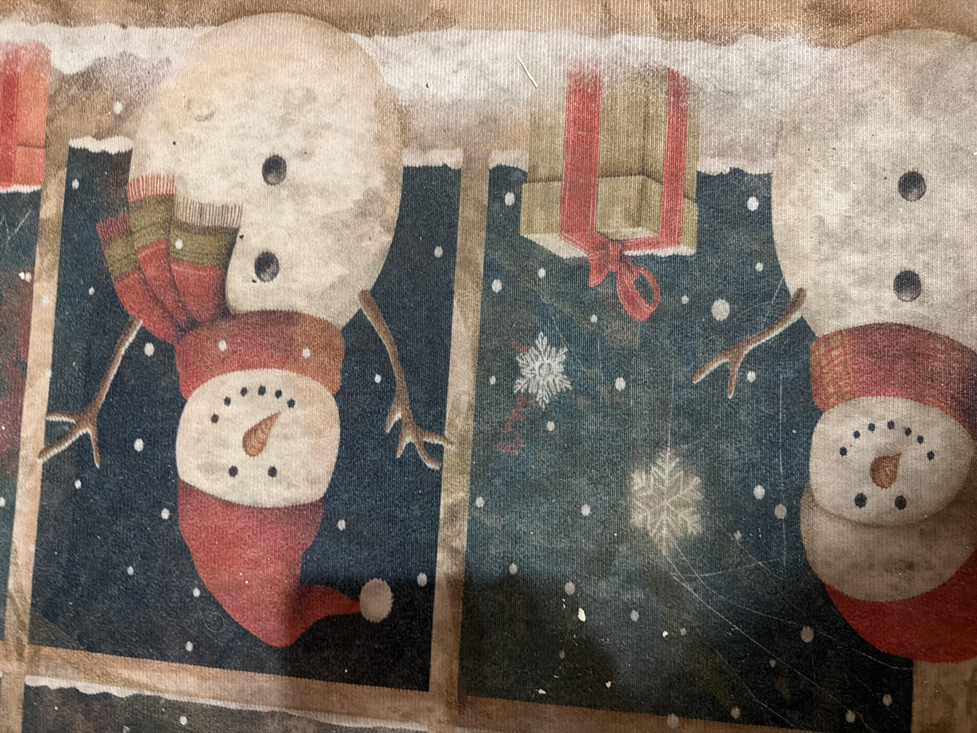 This image depicts a Christmas-themed artwork or textile, featuring two snowmen and holiday elements. The upper portion of the image shows a snowman wearing a red Santa hat and a striped scarf, with a carrot nose and button eyes. This snowman appears to be reaching out with a twig arm. Below this, a festive scene unfolds with snowflakes and what appears to be a stack of gift-wrapped presents tied with a red ribbon. The lower part of the image reveals another snowman, partially visible, also adorned with a red hat and scarf. The overall impression is of a wintry, cheerful holiday scene. There is no information in the image that allows for the identification of a specific location in the city of Armavir, Armenia.