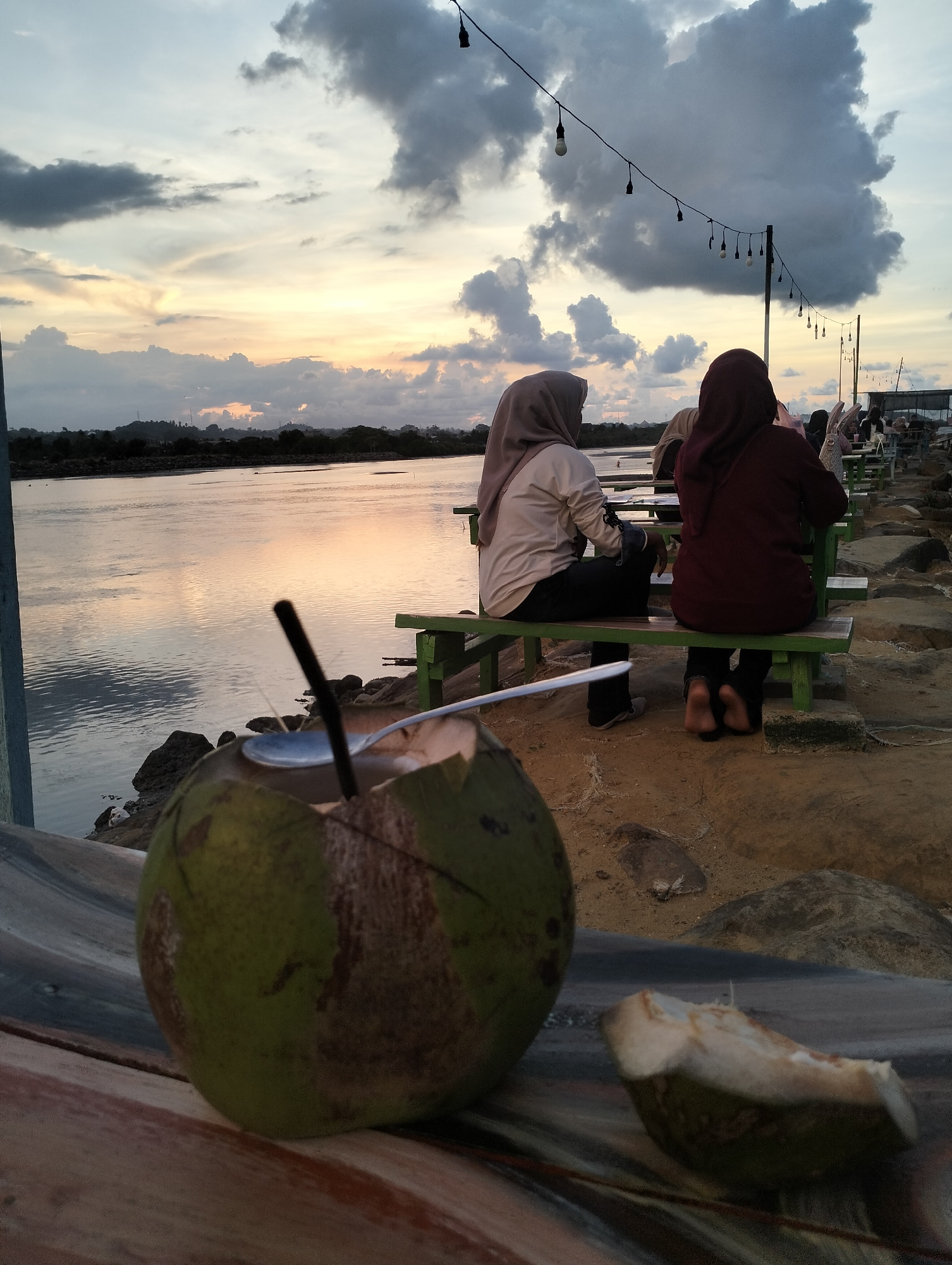 The photograph, taken in Lhokseumawe, Indonesia, presents a scene at dusk. In the foreground, a halved coconut with a straw and spoon rests on a weathered wooden surface. A piece of coconut shell lies next to it. Behind this, two women wearing hijabs sit on a green wooden bench along a waterfront. The woman on the left wears a light-colored shirt and dark pants, while the woman on the right wears a burgundy shirt and dark pants, with bare feet resting on the ground.
Further in the background, several other people are seated at similar benches, suggesting a public seating area or establishment. A body of water reflects the orange and gray hues of the setting sun and overcast sky. A string of lights hangs above the seating area, adding to the ambiance. The terrain appears to be rocky and sandy. The sky is a mix of dark clouds and lighter areas illuminated by the setting sun. Vegetation is visible in the far distance.