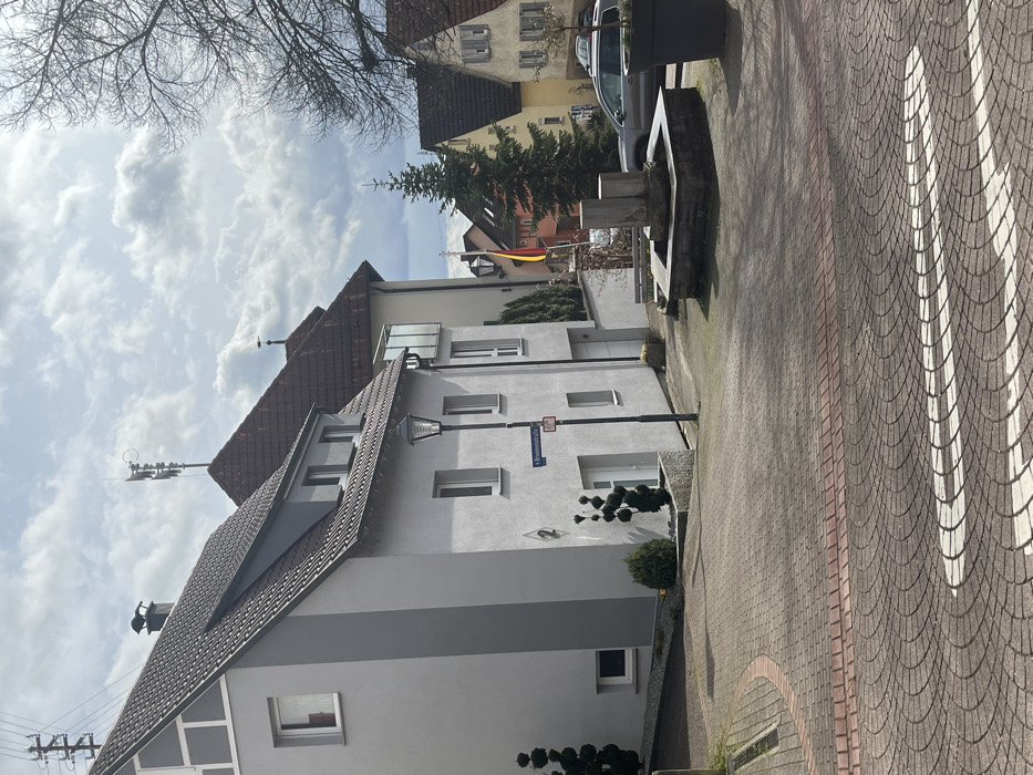 This image depicts a residential street scene in Hardthausen am Kocher, Germany, specifically looking towards Brunnenstraße 4 and the surrounding area.
The foreground features a paved surface composed of interlocking pavers, primarily in shades of reddish-brown and grey-brown, arranged in a repeating pattern. White, curved pedestrian markings are visible on this surface, along with a narrow strip of red brick pavers. To the far left, a metal grate drain is visible within the paving.
On the left side of the frame, a prominent two-story residential building, light grey in color with a dark brown tiled gabled roof, occupies the mid-ground. This building features multiple rectangular windows with white frames. A small dark grey dormer is present on the roof. Near the main entrance, a black house number "2" is affixed to the wall. Adjacent to the building, a street lamp with a black post and a multi-pane lantern head stands. Below the lamp, a blue street sign with white text "Brunnenstraße" and a red circular sign with a diagonal white bar are attached. A topiary shrub, shaped in a spiral, is planted in a grey planter near the building entrance, with a smaller green shrub next to it. Another similar topiary is visible further left.
Behind the initial grey building, another light-colored residential structure with a gabled roof and a balcony with glass railings is partially visible. Further back and to the right, additional residential buildings with varying rooflines and wall colors (some yellow, some with reddish-brown tiled roofs) are present. A German flag (black, red, gold) flies from a pole in the mid-ground, positioned between buildings. An evergreen conifer tree stands vertically to the right of the flag.
In the center-right of the image, a square-shaped stone fountain with a central vertical stone column structure is situated in a small paved area. No water is visible. A light-colored SUV is parked on the street further down, behind the fountain area. To the far right, a large, dark grey planter pot is visible, and the branches of a bare deciduous tree extend into the upper right corner of the frame. On the roof of a more distant building, a weather vane is visible, and on the roof of a building closer to the left, a multi-horn siren system is mounted. Overcast sky with scattered white clouds is visible in the background. No individuals are present in the scene.