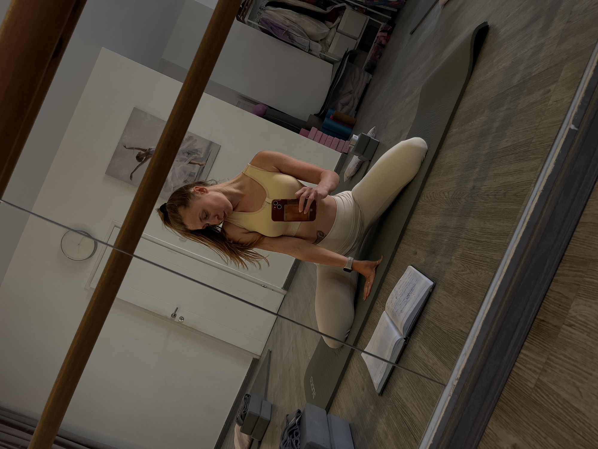 A mirrored view shows a woman in workout clothes holding a phone while kneeling on a yoga mat. The mat rests on a wooden floor. An open book sits next to her on the mat. Behind her are yoga blocks, and in the background, a clothing rack. A framed picture hangs on the wall. A clock is visible behind the clothing rack. The location appears to be a home or studio. There is no indication of the location being in Budva, Montenegro.
