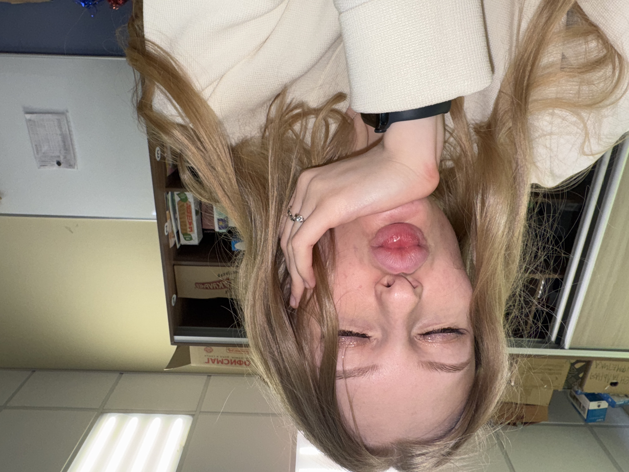 A young woman is upside down, posing for a selfie. She wears a light-colored coat and a watch. Her hair is light brown. She holds her hand near her face, and her lips are pursed. There appears to be a tear on her cheek. In the background, there's a wall-mounted cabinet and various boxes. The box labeled "ОФИСМАТ" may suggest the location is in Russia. The location of the photo, Perm, Russia, cannot be confirmed from this image alone.