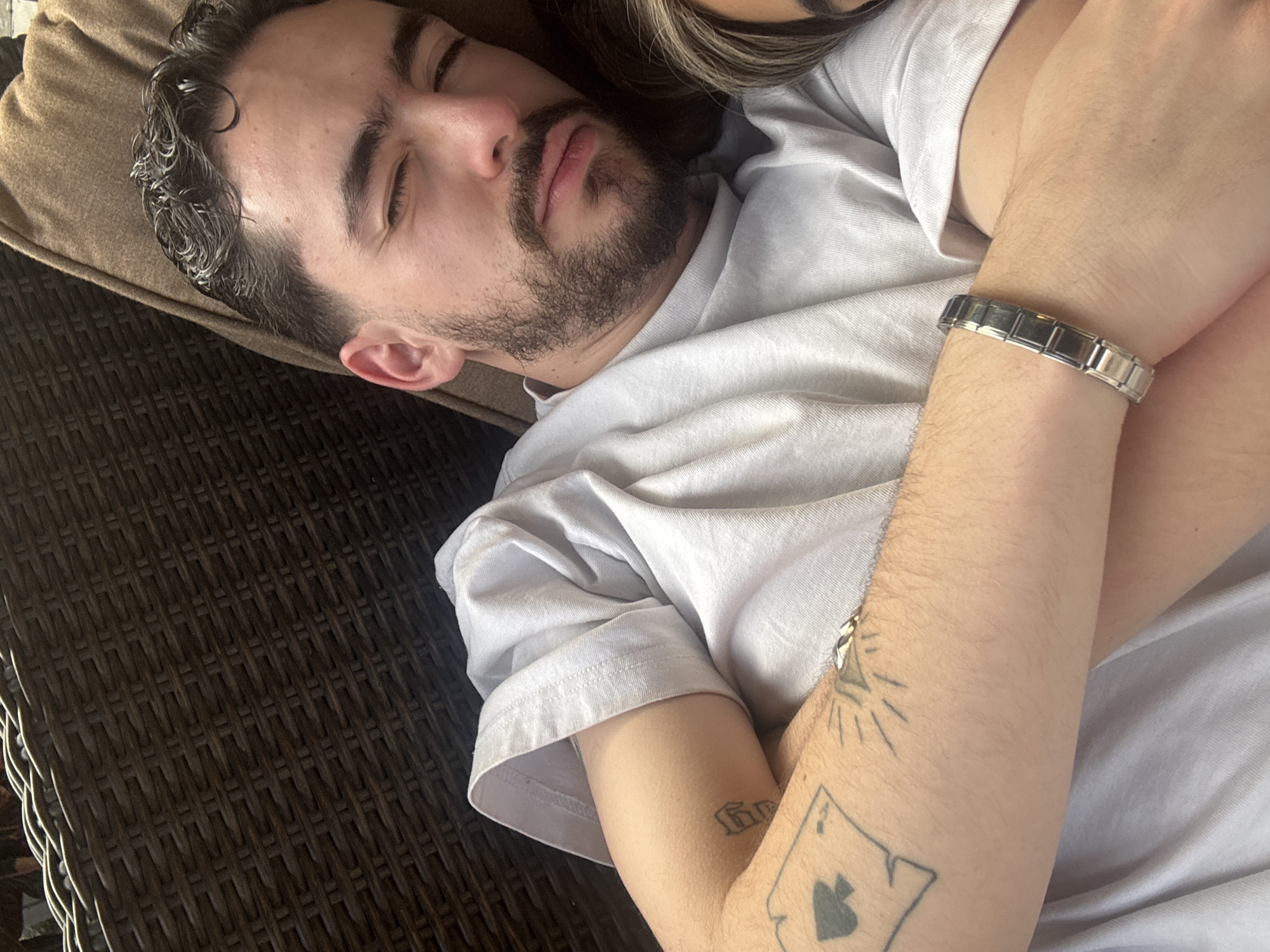 A man with a beard is lying down, possibly asleep. He is wearing a white t-shirt and has a silver bracelet on his wrist. Tattoos are visible on his arm. He is resting on a cushion that appears to be on a wicker or woven surface, possibly outdoor furniture. The location is Unknown, Unknown.