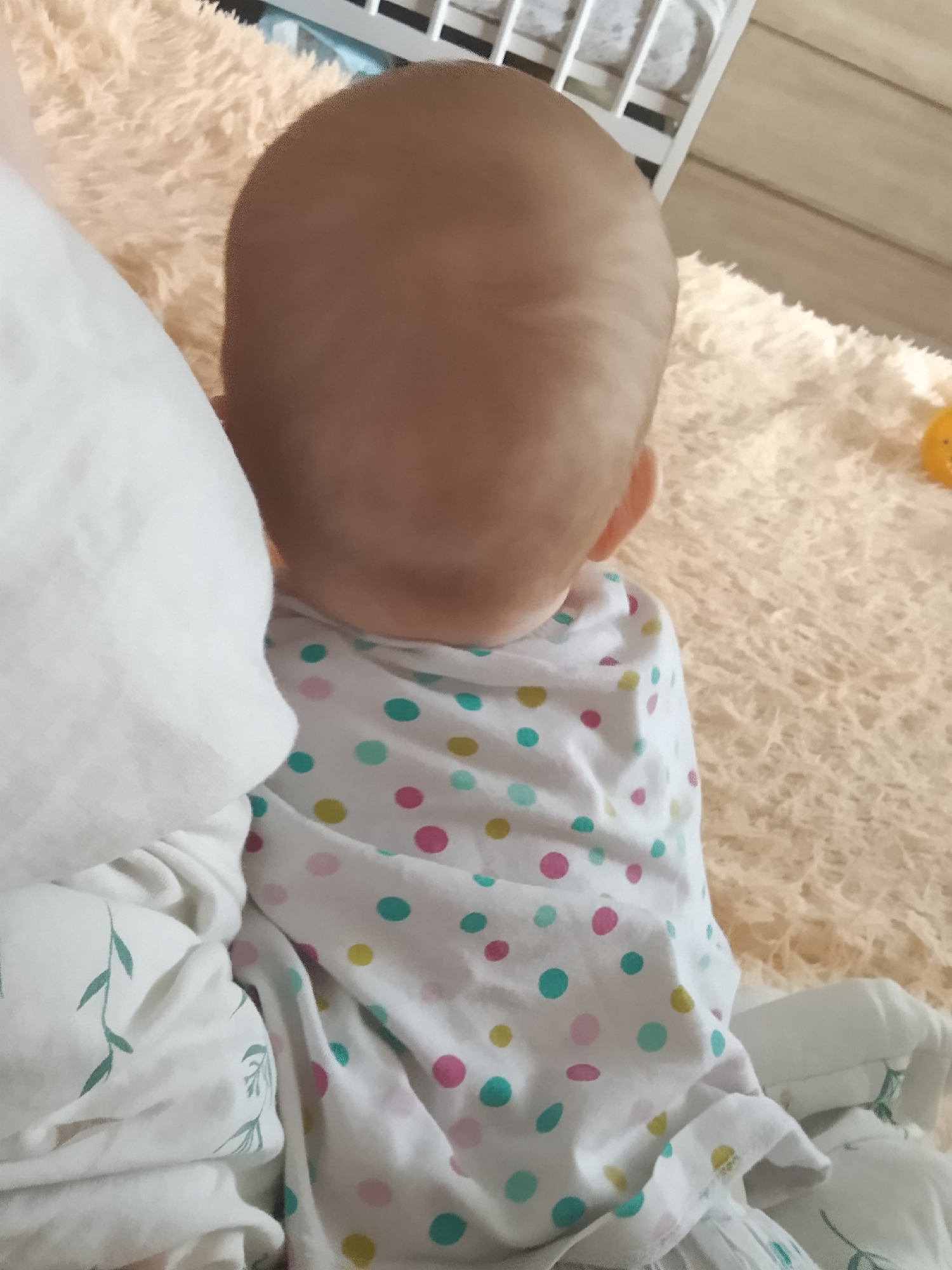The image shows the back of a baby's head and shoulders, wearing a white onesie with a polka dot pattern. The baby is lying on a soft, peach-colored furry blanket. To the left of the baby is a white cloth, possibly a blanket or a pillow, with a green vine pattern. In the background, a wooden surface and a white crib railing are visible. The scene appears to be indoors, in a domestic setting. There is no information or visual evidence in the image that suggests the location is Minsk, Belarus.