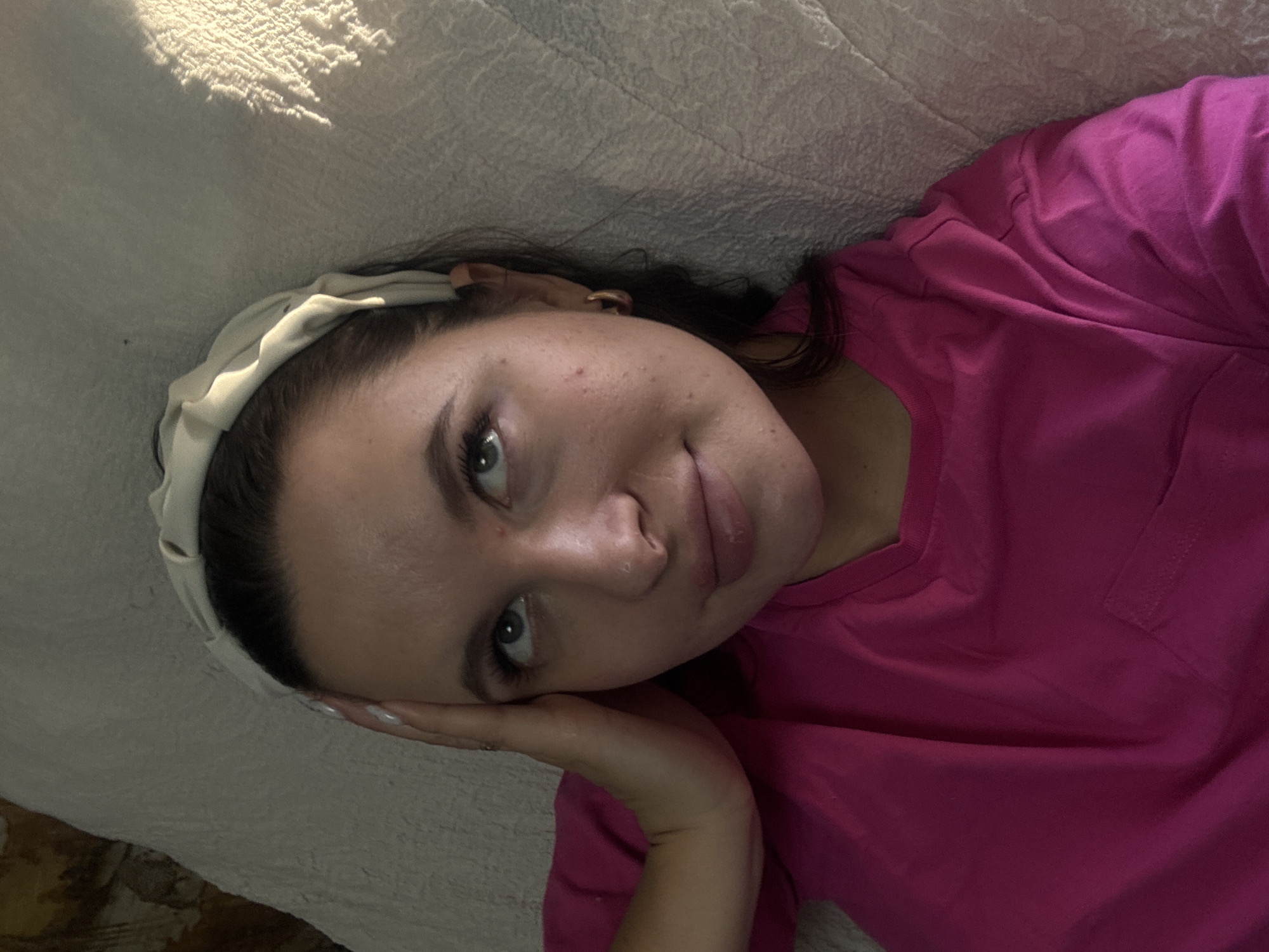 A woman lies on a textured, light-colored surface, possibly a bed or couch. She is wearing a pink short-sleeved shirt and a light-colored headband. Her dark hair is visible beneath the headband. She is resting her head on her hand, and her face is angled towards the camera. Her eyes are open and she has fair skin with slight blemishes. The image has soft, natural lighting. The location, Красный Маяк, Russia, is not visually discernible in the image.