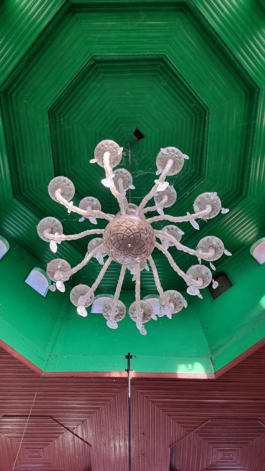 An ornate, multi-tiered chandelier hangs from a vibrant green, octagonal ceiling in what appears to be an interior space. The chandelier features numerous clear, faceted glass elements and a central, textured glass orb. Subtle cobwebs are visible clinging to the chandelier and the ceiling. The ceiling is constructed with wooden planks arranged in a geometric pattern. Below the green ceiling, a section of dark reddish-brown wooden paneling with a similar geometric design is visible, suggesting the structure of the room. In the lower portion of the image, a tall, slender black pole with a small attachment at the top extends upwards towards the chandelier, possibly a light stand or part of an overhead fixture. The overall scene suggests an indoor architectural setting with decorative lighting. The location is identified as Blangrambong, Indonesia, though the specific context within Blangrambong is not discernible from the image alone.