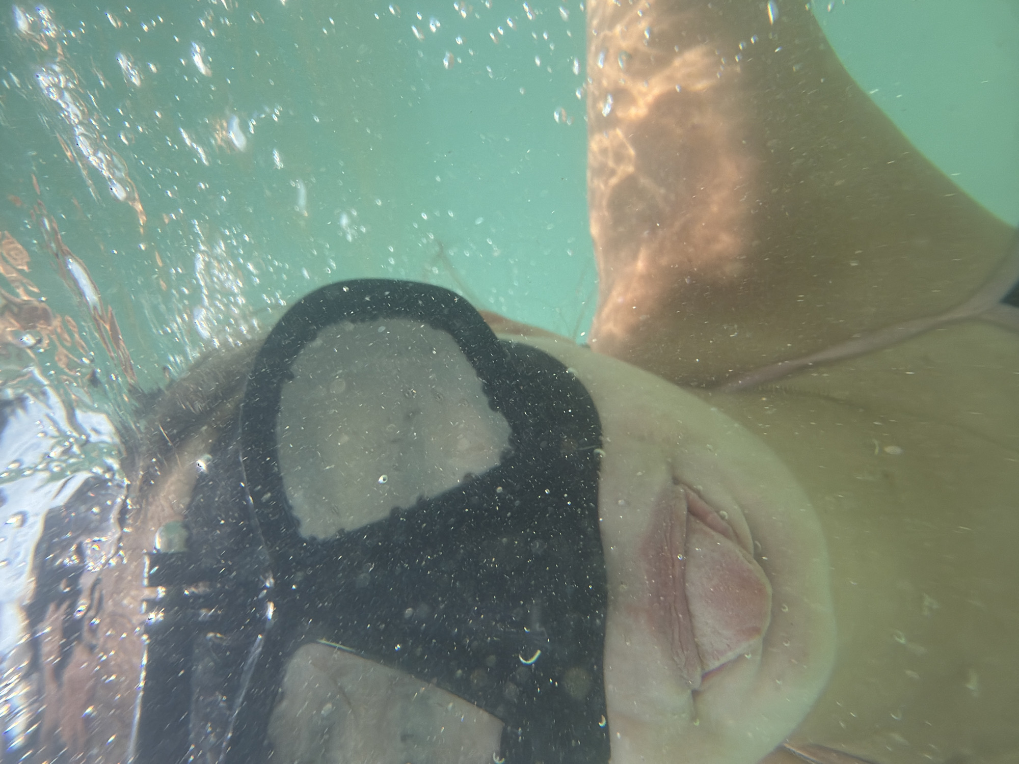 An extreme close-up underwater shot captures a person's face, partially submerged in clear turquoise water. The subject wears a black diving mask that covers their eyes. Their mouth is open, with their tongue extended slightly. Bubbles are visible throughout the water, indicating movement and aeration. The lighting suggests it is daytime, with sunlight filtering through the water, creating dappled patterns on the subject's skin. The overall environment appears to be a natural body of water, such as the ocean, and the presence of clear water and sunlight is consistent with the coastal environment of Nha Trang, Vietnam.