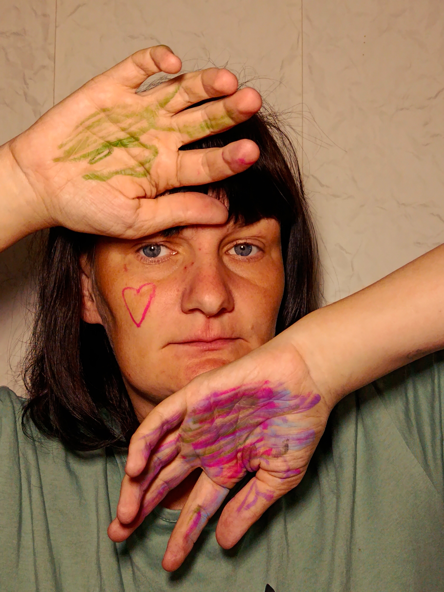 A fair-skinned woman with dark hair and blue eyes faces the camera. She is wearing a light green t-shirt. Her hands are raised, with one hand above her forehead and the other obscuring the side of her face. Her palms are covered in green and purple/pink paint. A pink heart shape is drawn on her left cheek. The background is a light beige wall. There is no indication of Фоки, Russia.