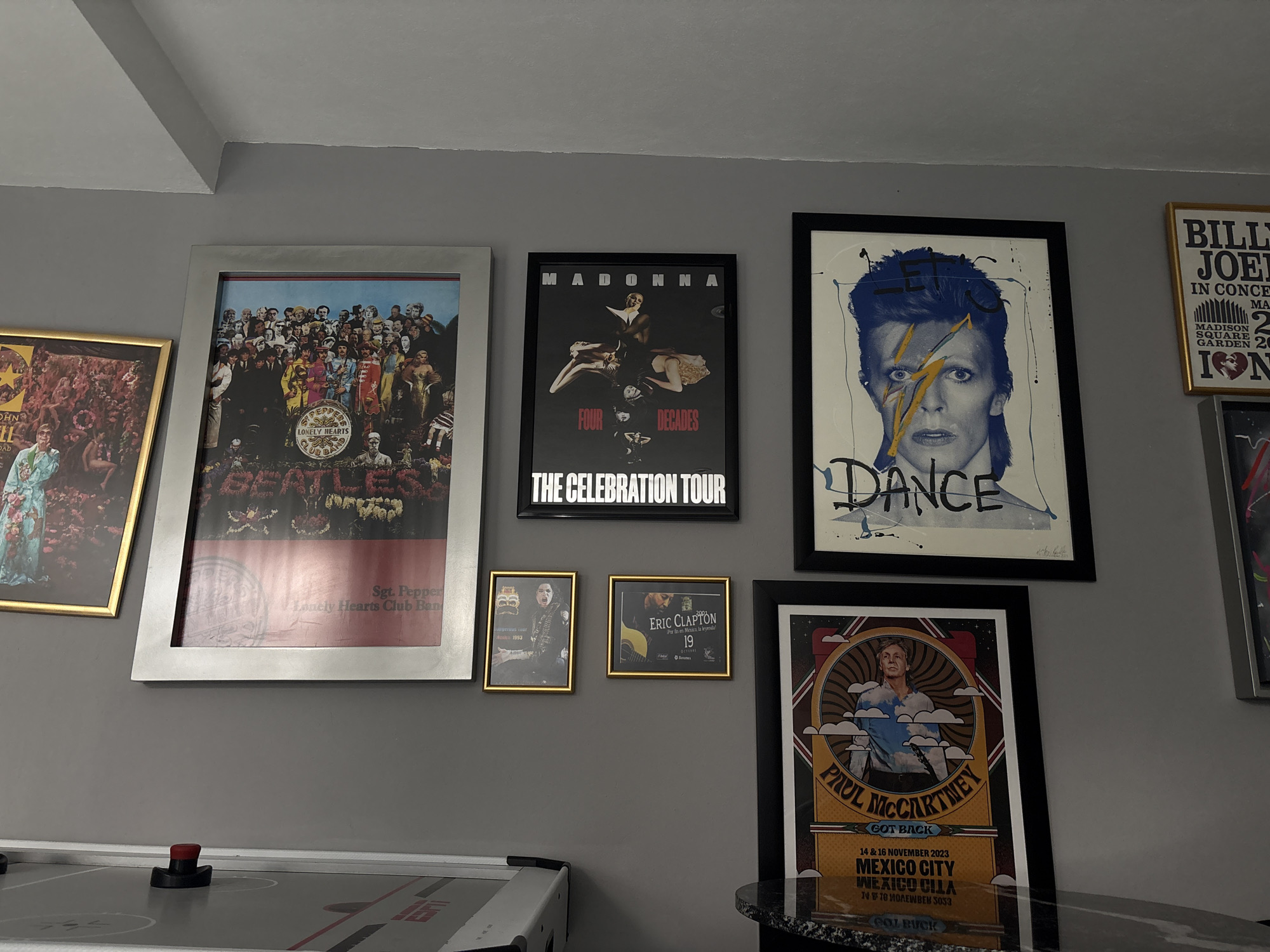 A collection of framed posters and memorabilia is displayed on a gray wall. The posters are arranged in a somewhat asymmetrical grid, with the largest poster on the left featuring The Beatles' "Sgt. Pepper's Lonely Hearts Club Band" album cover. To its right is a Madonna "The Celebration Tour" poster. Adjacent to that is a David Bowie "Let's Dance" poster. Below these are smaller framed posters, including Eric Clapton and Paul McCartney "Got Back" Mexico City, 14 & 16 November 2023. A portion of an air hockey table is visible at the bottom left of the frame. San Bernardino Tlaxcalancingo is not recognizable in the image.
