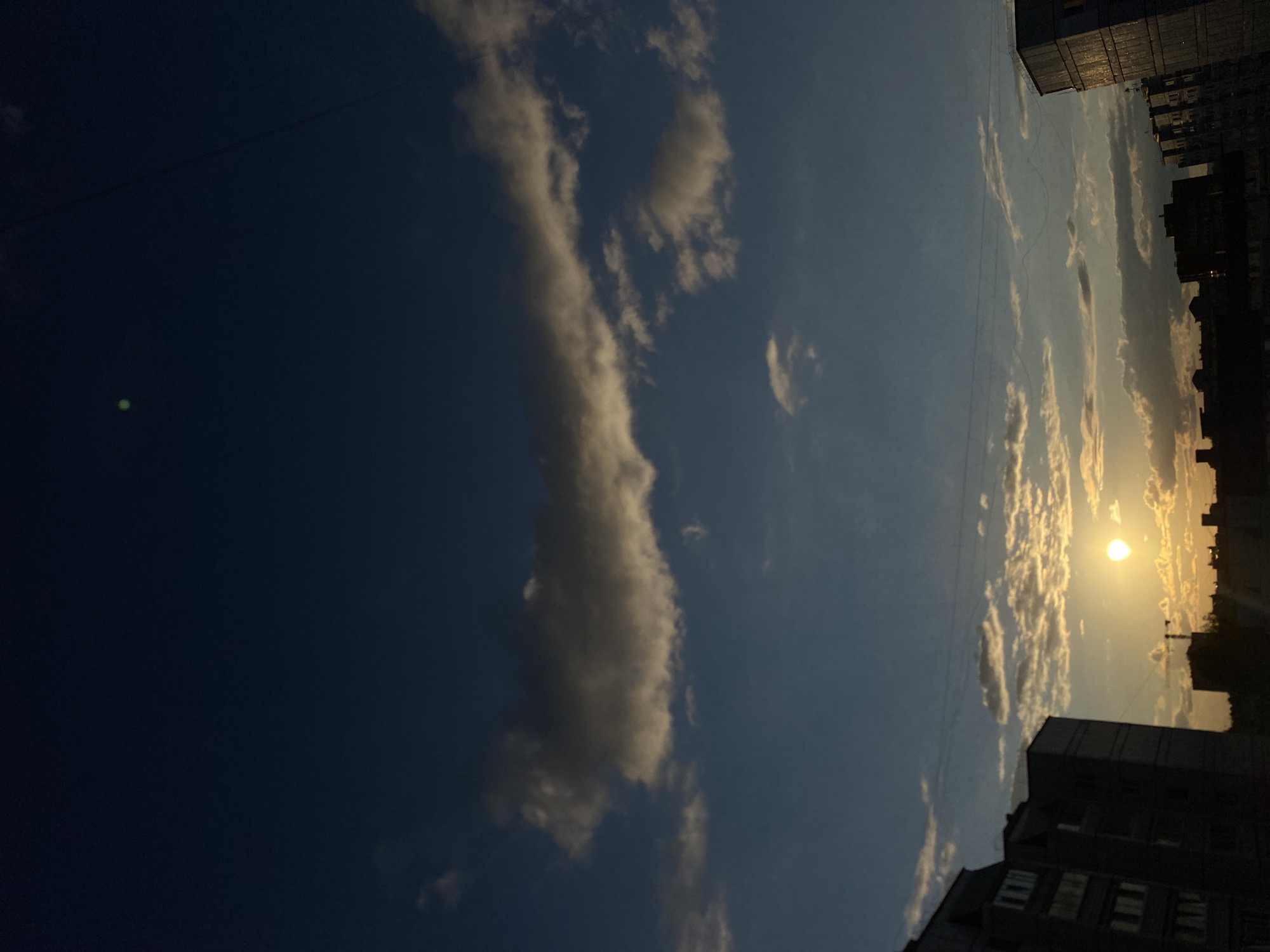This image shows a sunset over the city of Chaikovsky, Russia. The sky is a deep blue, with streaks of orange and yellow from the setting sun. Clouds are scattered across the sky, some dark and ominous, others light and wispy. In the foreground, silhouetted buildings rise against the colorful sky. The buildings appear to be apartment blocks, typical of Soviet-era architecture. A construction crane is visible in the distance. The overall impression is one of urban tranquility, with the beauty of nature juxtaposed against the man-made environment.