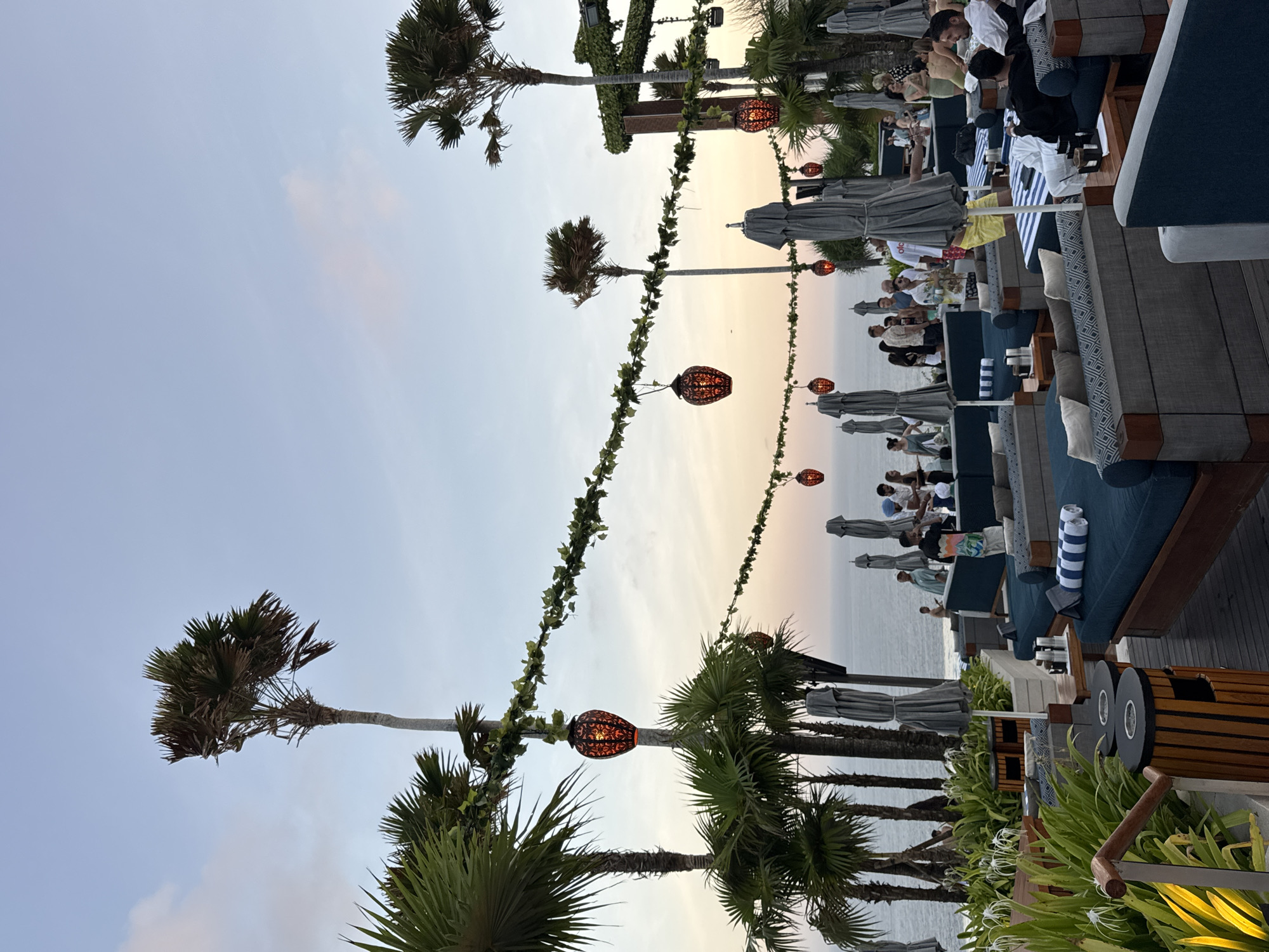 This media file captures an outdoor dining scene at dusk, likely in Kuta Selatan, Indonesia. The setting appears to be a stylish beach club or restaurant with a view of the ocean.
The primary subjects are a group of people seated at tables, engaged in conversation and enjoying their evening. They are dressed in casual attire, suggesting a relaxed atmosphere. The tables are set with striped napkins and some have drinks on them. There are also lounge chairs with blue cushions and umbrellas, indicating a comfortable seating arrangement.
Decorative elements include string lights with ornate lanterns hanging from palm trees, adding a warm ambiance to the scene as the sky transitions through shades of orange and pale blue. Several palm trees are prominent, their fronds reaching towards the sky. The ocean is visible in the background, and the weather appears calm and pleasant.
The time of day is clearly evening, with the warm glow of the sunset. The overall mood is one of leisure and enjoyment, typical of a vacation destination. There is no visible text in the image.