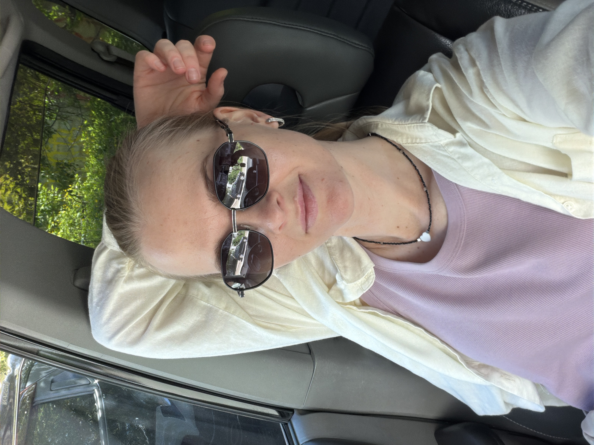 The image is a selfie taken from the perspective of someone sitting in the passenger seat of a car, looking towards the driver. The woman has blonde hair pulled back and is wearing sunglasses, a light-colored open button-down shirt over a lavender tank top, and a beaded necklace with a white pendant. Her left arm is resting on the headrest behind her. The interior of the car is visible, including a dark gray leather seat. Through the car windows, foliage and a building with yellow graffiti are visible, suggesting an outdoor setting, possibly in a residential area or park. Based on the visual cues such as typical Russian car models (e.g., the visible rear of a black SUV on the left) and architectural styles, as well as the specific type of graffiti, it is plausible this scene is located in Khabarovsk, Russia, likely during the warmer months given the foliage.