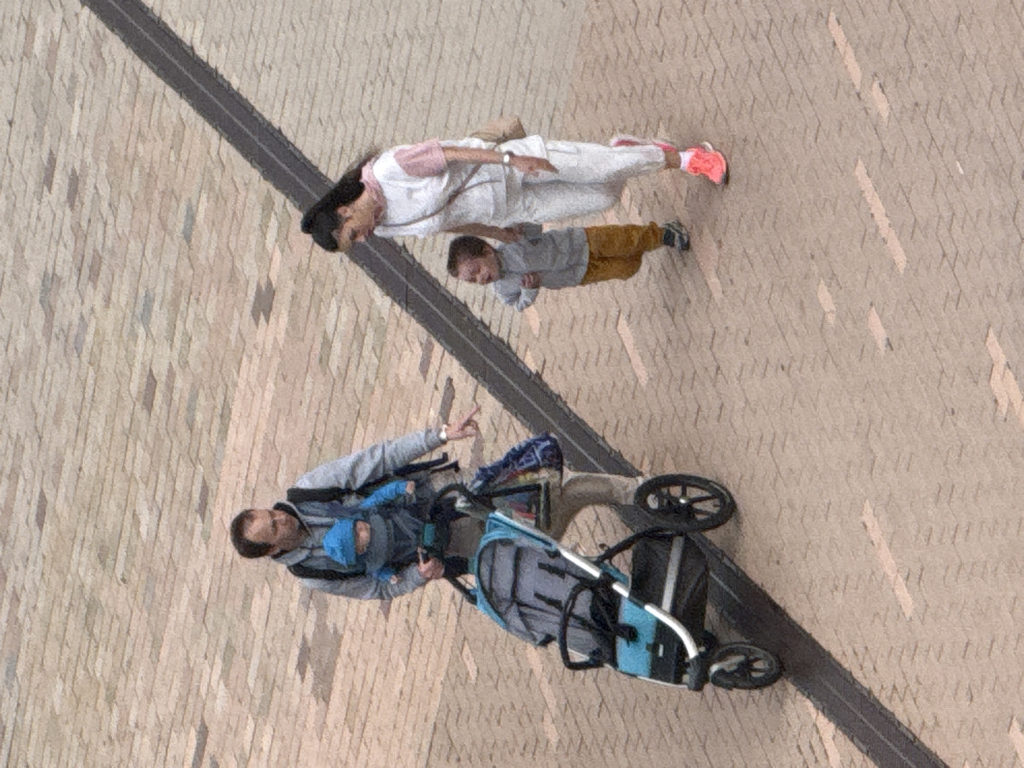 A family of four is walking on a paved pedestrian area with a textured surface. A man in a grey hoodie, holding a baby in a blue carrier on his chest, pushes a blue and grey stroller. To the right of the man, a woman in white pants and a pink and white top walks with a young child in a grey jacket and orange pants. The child is holding the woman's hand. The area appears to be outdoors, possibly a park or public square, with a dark, diagonal strip of material set into the paving. The lighting suggests it is daytime. This scene is located in Khimki, Russia.