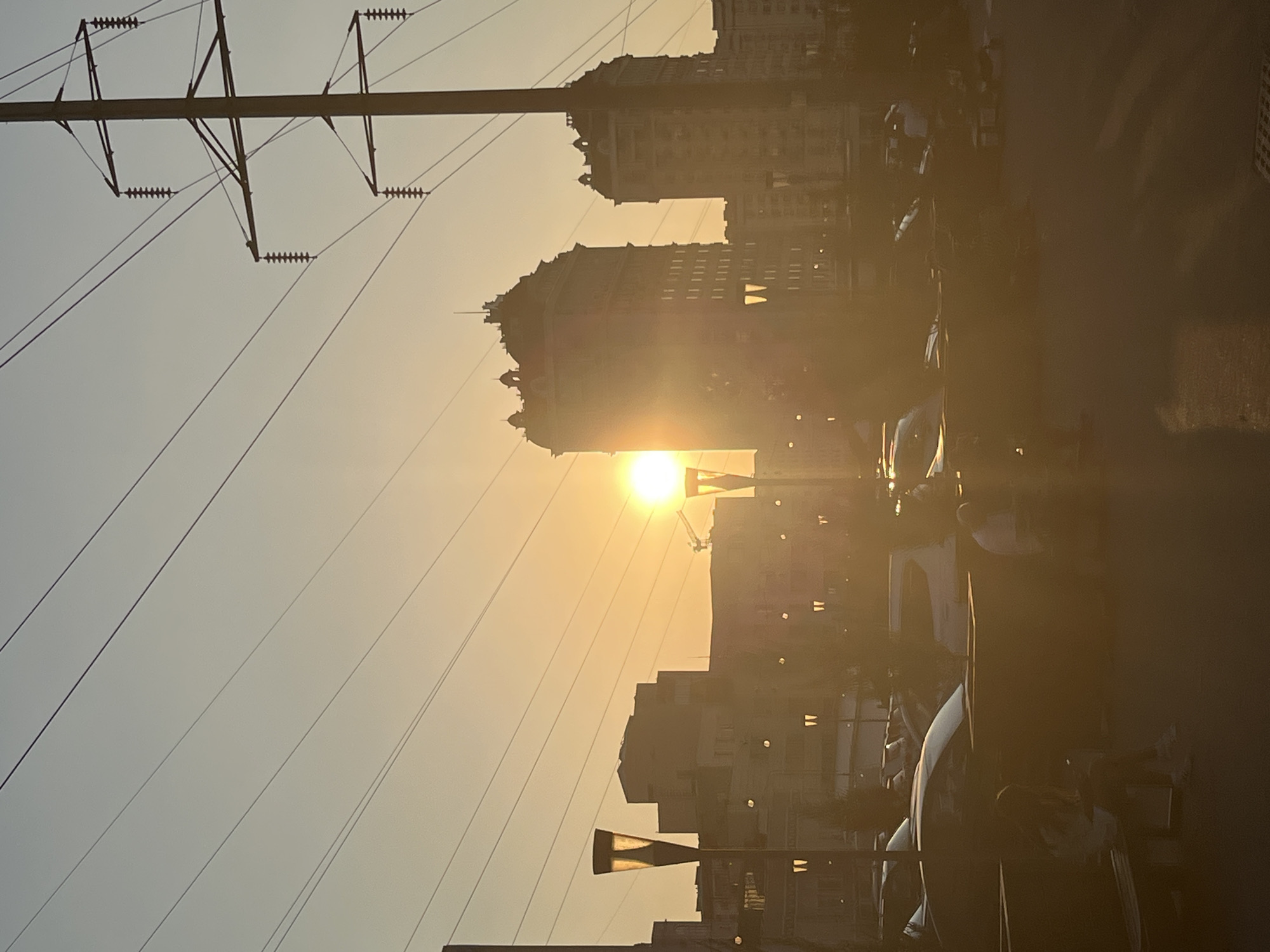 This image depicts a sunset over Bishkek City, Kyrgyzstan. The sun, a bright orb, is positioned in the middle left of the frame, casting a warm glow across the scene. Silhouetted against the sky are several tall buildings, some with ornate architectural details, and a prominent electrical pylon with power lines stretching across the frame. In the foreground, streetlights with distinctive lamps illuminate the area, casting pools of light. A parking lot with several cars is visible, and in the lower portion of the image, there are blurred figures of people sitting on benches. The overall impression is a serene urban landscape at dusk, bathed in the golden light of the setting sun.