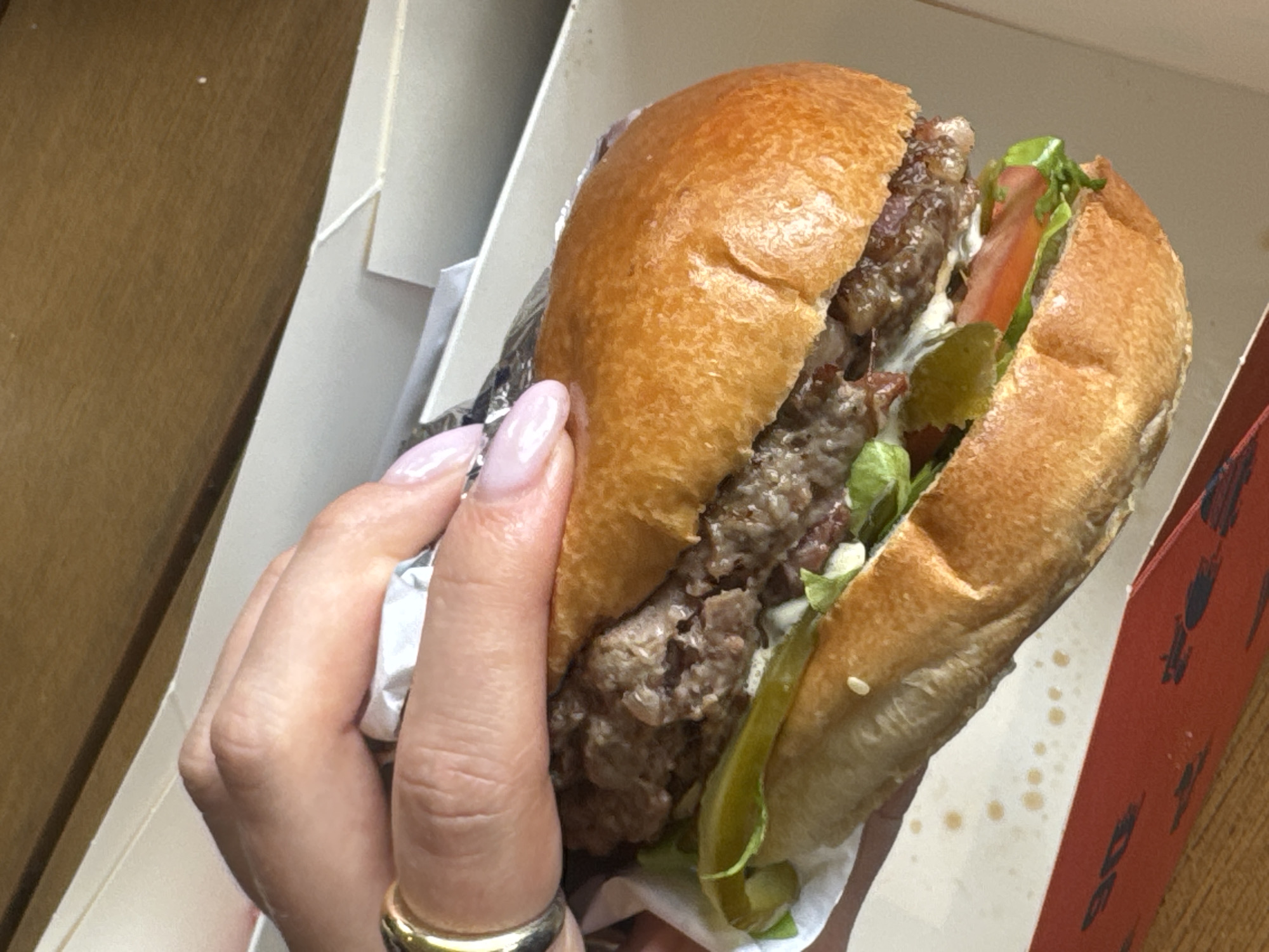 A partially eaten hamburger held in a person's hand is positioned inside a white cardboard container with red design elements. The hamburger consists of a browned bun, a beef patty with visible texture, lettuce, sliced tomato, pickle slices, and what appears to be a white sauce or mayonnaise. The person's hand is visible, featuring pink nails and a gold ring. The container is slightly soiled with grease stains. The background suggests a wooden surface and interior setting in the city of Unknown, Unknown.