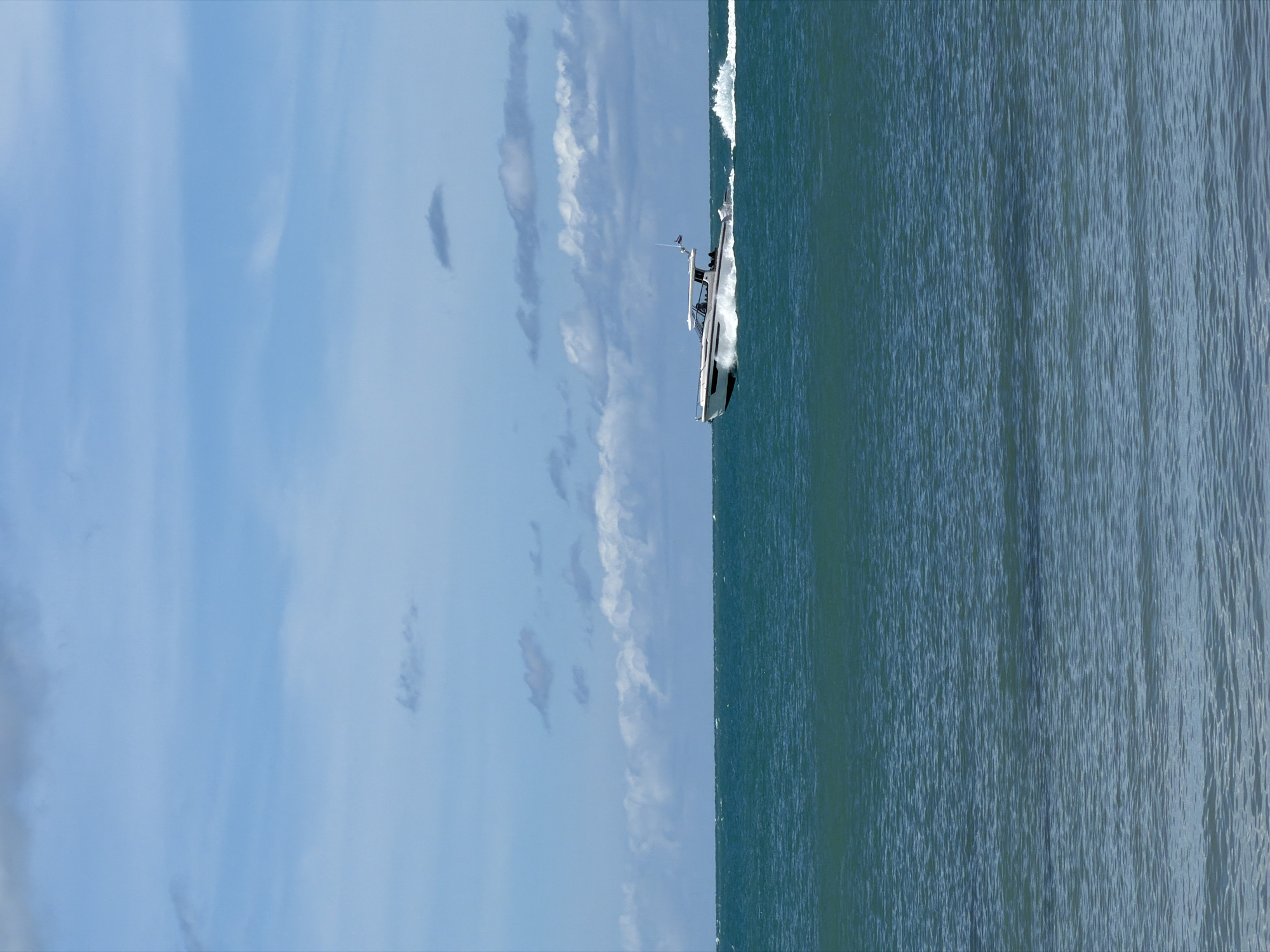 The image shows a white motorboat with a black hull speeding across choppy blue water. The boat is angled diagonally in the frame, with its bow pointed towards the upper right corner. There are several people visible on the boat, appearing to be passengers. The boat is creating a white wake behind it.
The scene is set against a backdrop of a vast, pale blue sky with scattered, wispy white clouds. The water occupies the right side of the frame, showing a textured surface with many small ripples and waves. The lighting suggests it is daytime, likely with bright sunlight, though the exact time is difficult to determine. The location is identified as Baan Plai Laem, Thailand, implying a coastal or open sea environment. No text is visible in the image.