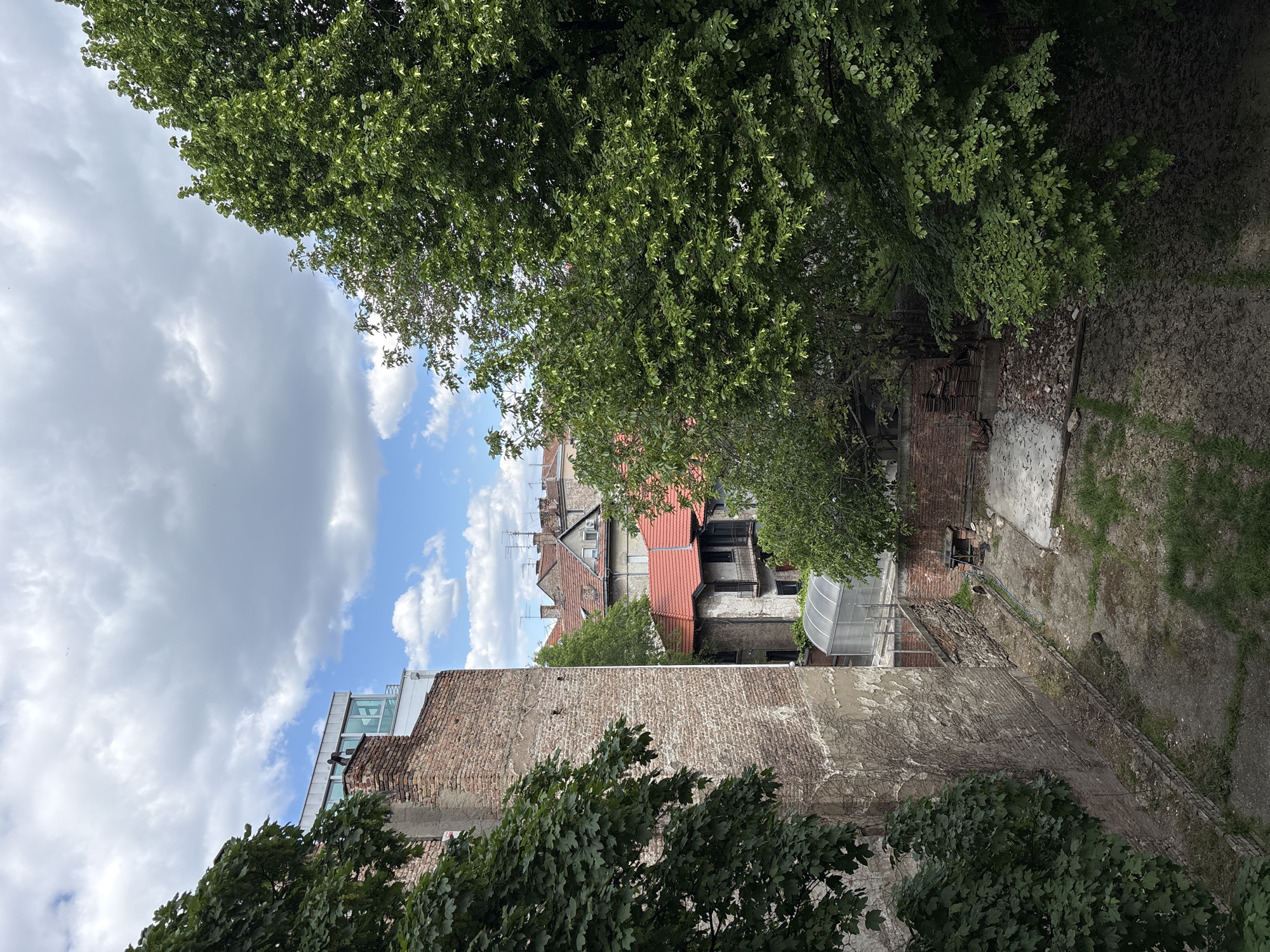 This is an urban outdoor scene, likely viewed from an elevated position overlooking a city courtyard or a gap between buildings. The image captures a vertical perspective of the scene, with the sky at the top and ground at the bottom if viewed in its natural portrait orientation.
In the foreground, a prominent, weathered brick wall runs diagonally, partially covered by lush green foliage, possibly ivy or creepers. Dense tree canopies, some with variegated leaves, are also visible in the lower-left and upper-right areas, framing the view.
The midground features an unpaved, somewhat unkempt ground area, comprising dirt, patchy grass, and scattered debris and rubble near the base of the walls. Further back, another brick wall defines the boundary of this space.
In the background, a cluster of diverse urban architecture rises. Several traditional residential buildings with pitched red-tiled roofs, chimneys, and external air conditioning units or satellite dishes are visible. Their facades vary, suggesting different ages and conditions. Contrasting with these older structures, a modern building with large glass windows and a flat roof can be seen in the upper-left portion of the background. An interesting detail is a transparent, arched roof structure, possibly a greenhouse or conservatory, attached to one of the older buildings.
The upper-left section of the sky, above the buildings and trees, is filled with a mix of grey clouds and patches of clear blue sky. The lighting indicates it is daytime. The overall scene, devoid of human activity, presents a typical urban vista blending historic architecture with newer elements, characteristic of cities like Belgrade, Serbia. There is no visible text in the image.
