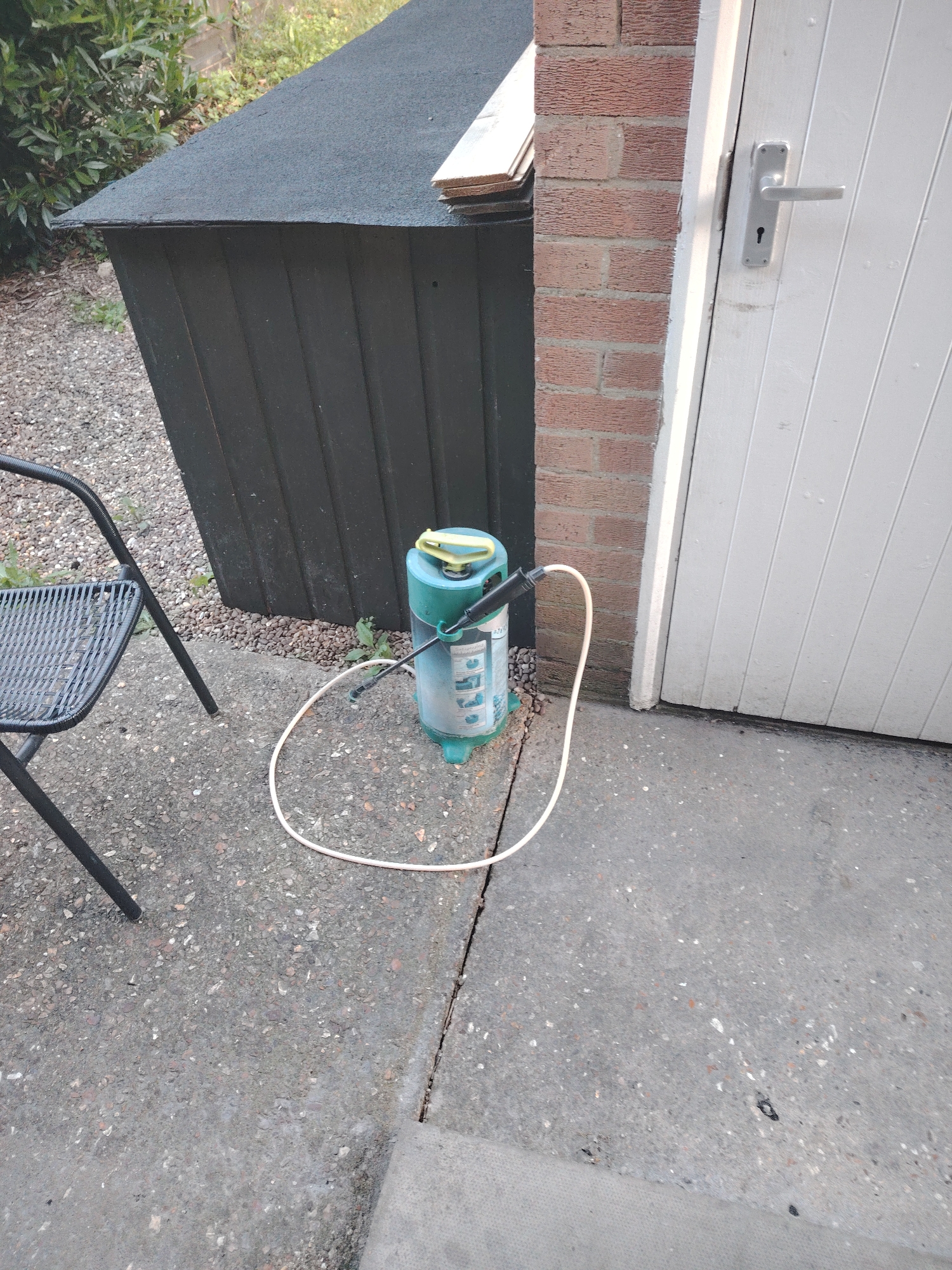 The image shows an outdoor scene in Huntingdonshire, United Kingdom. In the foreground, there is a concrete patio area with a crack running through it. A black metal chair with a woven seat is positioned on the patio. To the right of the chair is a green and blue pressure sprayer with a white hose. Behind the patio, there is a dark-colored wooden shed with a black felt roof. To the right of the shed is a brick wall and a white garage door with a silver handle. Overgrown green foliage is visible in the background to the left of the shed. The scene appears to be a domestic setting, possibly a garden or backyard.