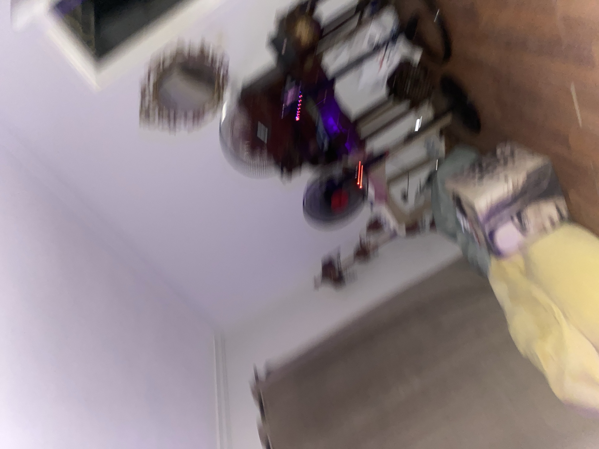 The image depicts an interior scene with an abstract and blurred aesthetic due to motion. In the foreground, a yellow object, possibly a blanket or clothing item, is partially visible. To the left, a tall, textured brown surface, likely a curtain or wall covering, dominates the frame. In the background, several objects are discernible, including what appear to be fans, one with a dark base and a red light. A framed mirror with an ornate, distressed frame hangs on a light-colored wall. The lighting is low, with purple hues contributing to the overall blurry and atmospheric quality. The setting suggests a domestic interior, but the lack of clear detail and the motion blur make precise identification difficult. The image is captured from a low angle, looking upwards towards the wall and ceiling. There is no indication of the specific location within Legenda, Egypt, or any activities taking place beyond the static presence of the objects.