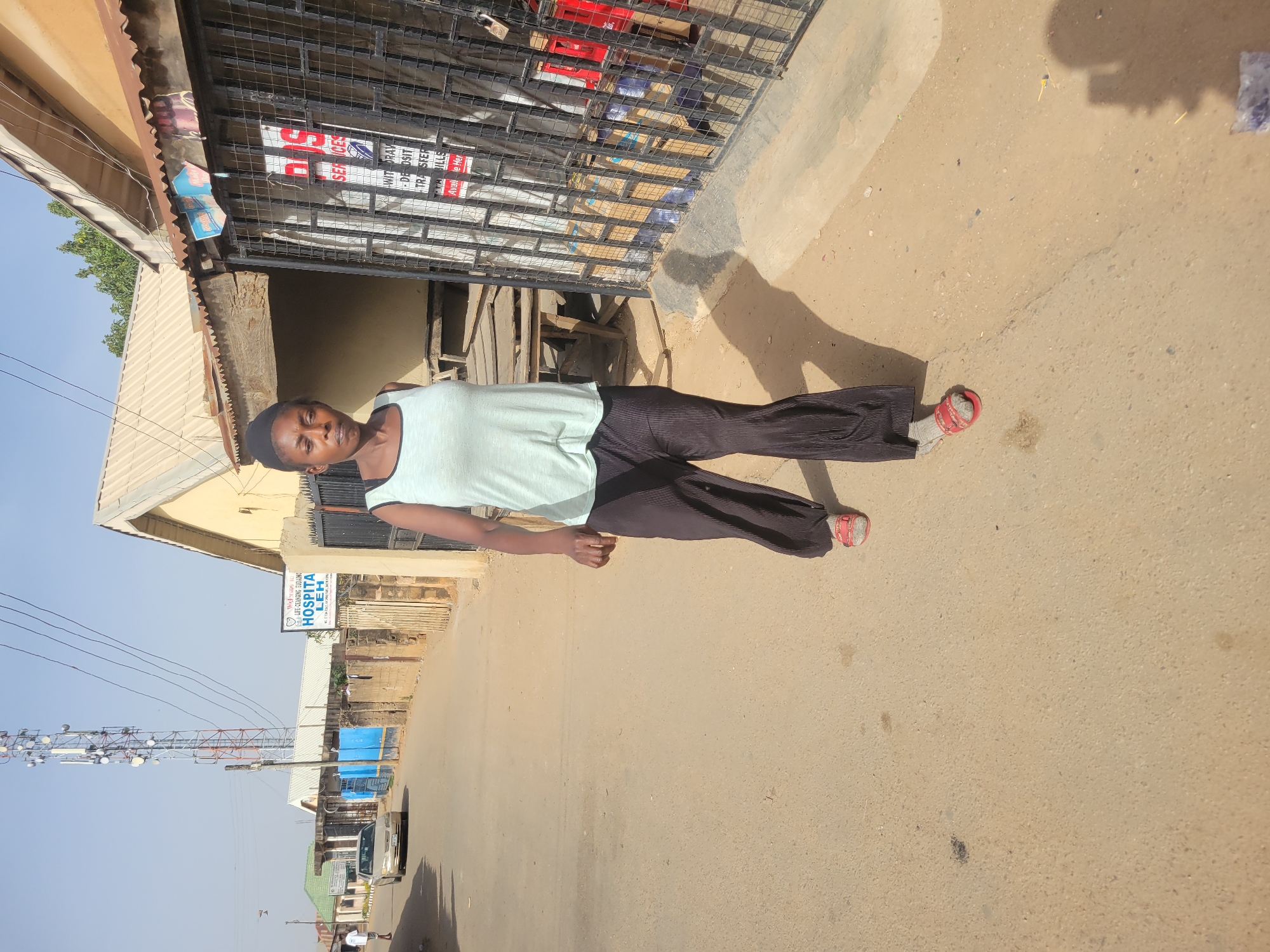 A person stands on a tan-colored road in what appears to be Jos, Nigeria. The individual wears a light-colored tank top and dark-colored, pleated pants, along with red sandals. They stand in front of a building with a corrugated metal roof and a barred gate. A sign reading "HOSPITAL LEH" is visible further down the street, where other buildings and a utility pole are also present. The sky is clear and blue.