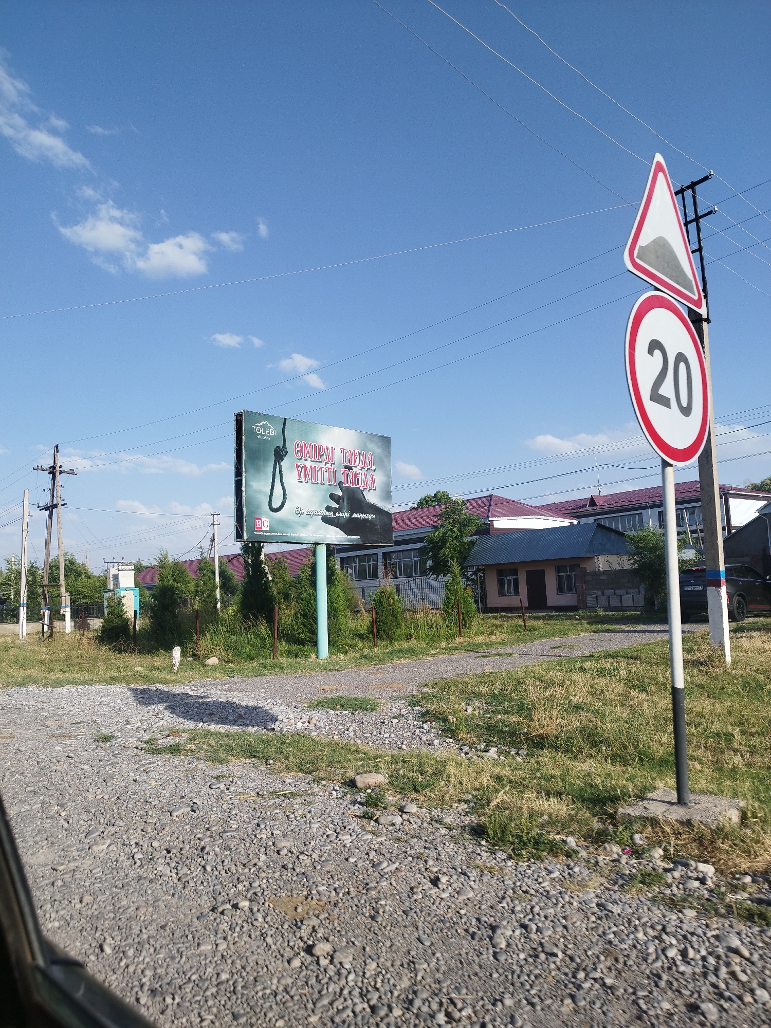 This is a daytime, eye-level, outdoor photograph taken in the Зертас ауылдық округі, Kazakhstan. The scene depicts a rural or suburban area with a gravel road in the foreground. To the right of the road, a traffic signpost displays two signs: a triangular warning sign with a red border and a symbol of a steep incline, and a circular speed limit sign indicating 20. Behind the signpost, a dark-colored car is parked. In the middle ground, a large billboard with a dark, dramatic advertisement is visible. The billboard shows a rope noose and text in an unknown language, with the brand name "TOLEBI" prominently displayed. Further back, a long building with a red roof and a smaller, light-colored building with a blue roof are situated. The background consists of a clear blue sky with scattered white clouds and power lines crisscrossing the frame. The vegetation in the image includes dry grass and several coniferous trees. The overall impression is one of a somewhat desolate, yet functional, roadside environment.