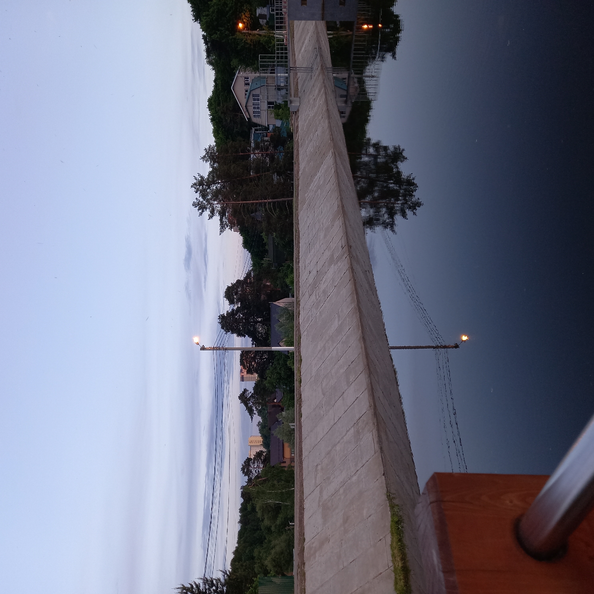 The image captures an evening scene near a body of water in Садовод-сад-3 Снт, Russia. A concrete retaining wall runs along the edge of the water, with reflections of trees, buildings, and streetlights visible on the dark surface. On the opposite side of the wall, there's a landscape with trees and buildings, including a multi-story house. Power lines are strung across the sky, connecting to streetlights. The sky is a gradient of light blue to dusky grey, suggesting twilight. A wooden railing is visible in the foreground, adding a sense of perspective.